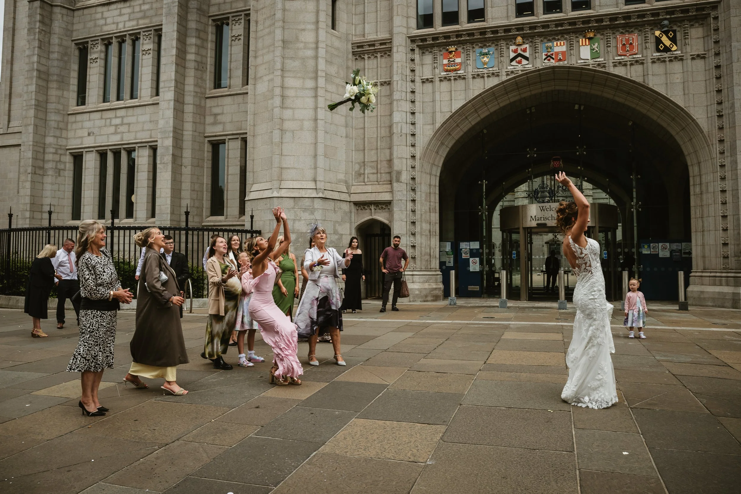 A bride in a white wedding dress throws her bouquet towards a group of women outside a large stone building with coats of arms.
