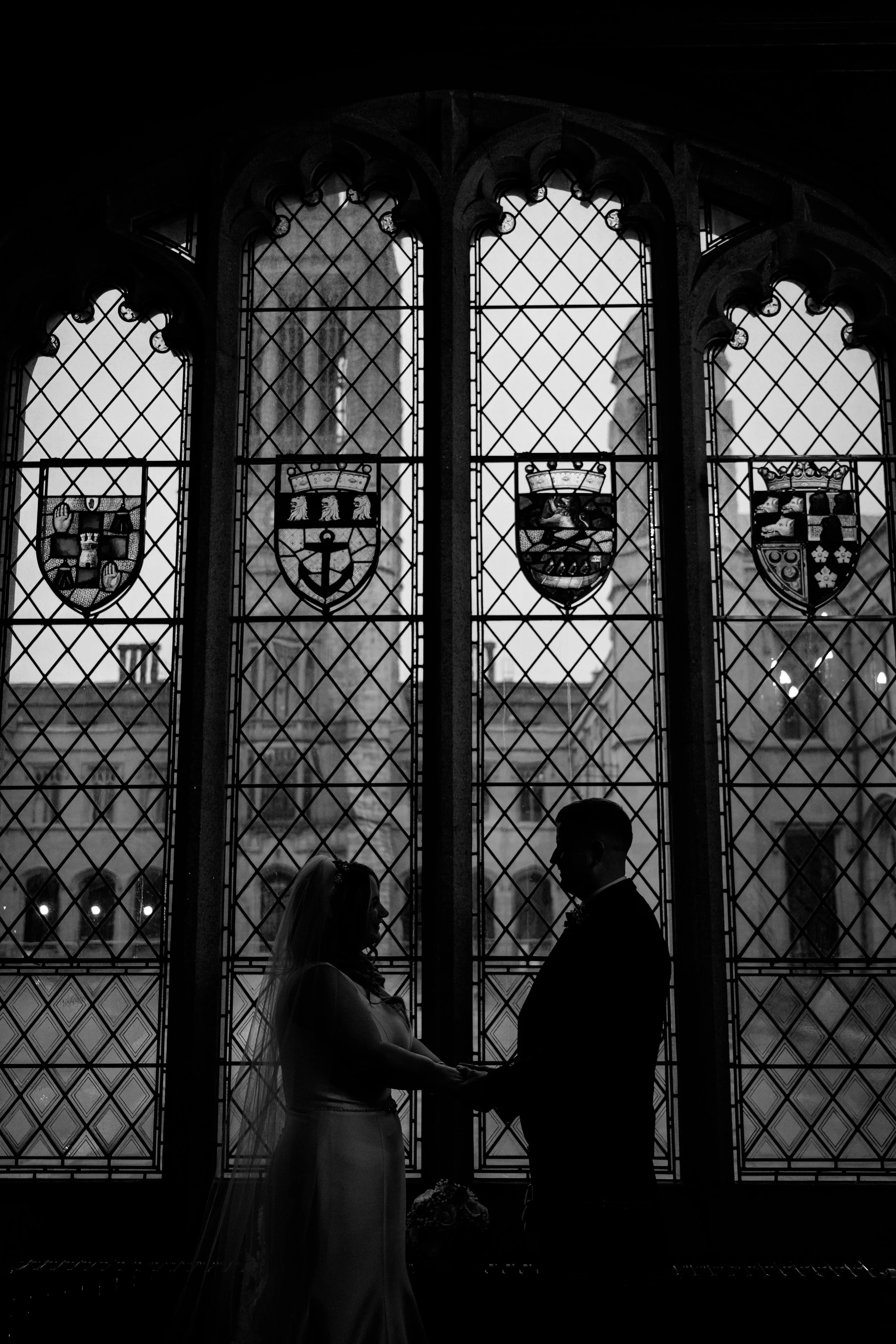 Silhouette of a bride and groom holding hands in front of tall stained glass windows with coats of arms, inside a church.