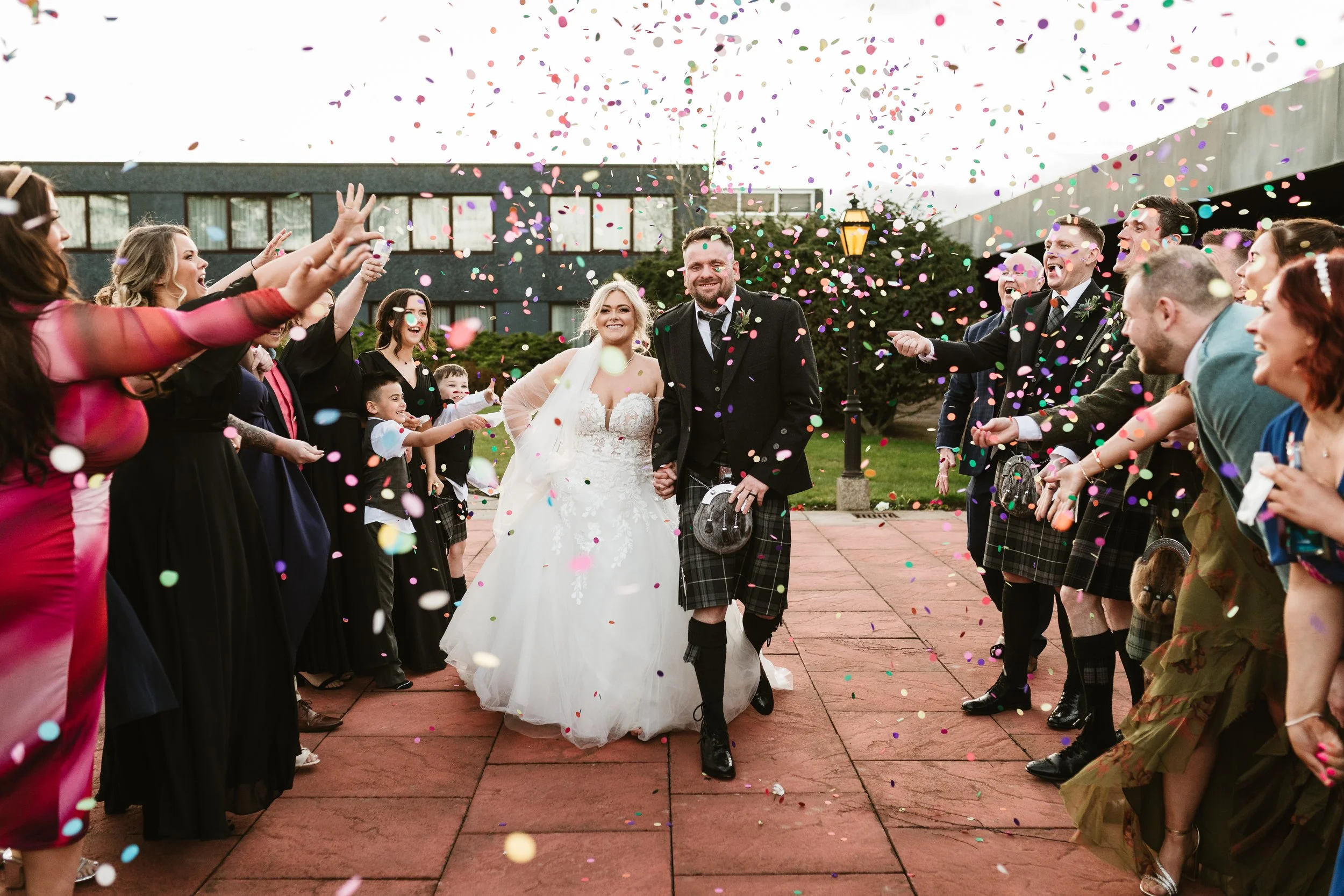 Bride and groom walking hand in hand through a crowd of wedding guests throwing confetti, outdoor wedding celebration.