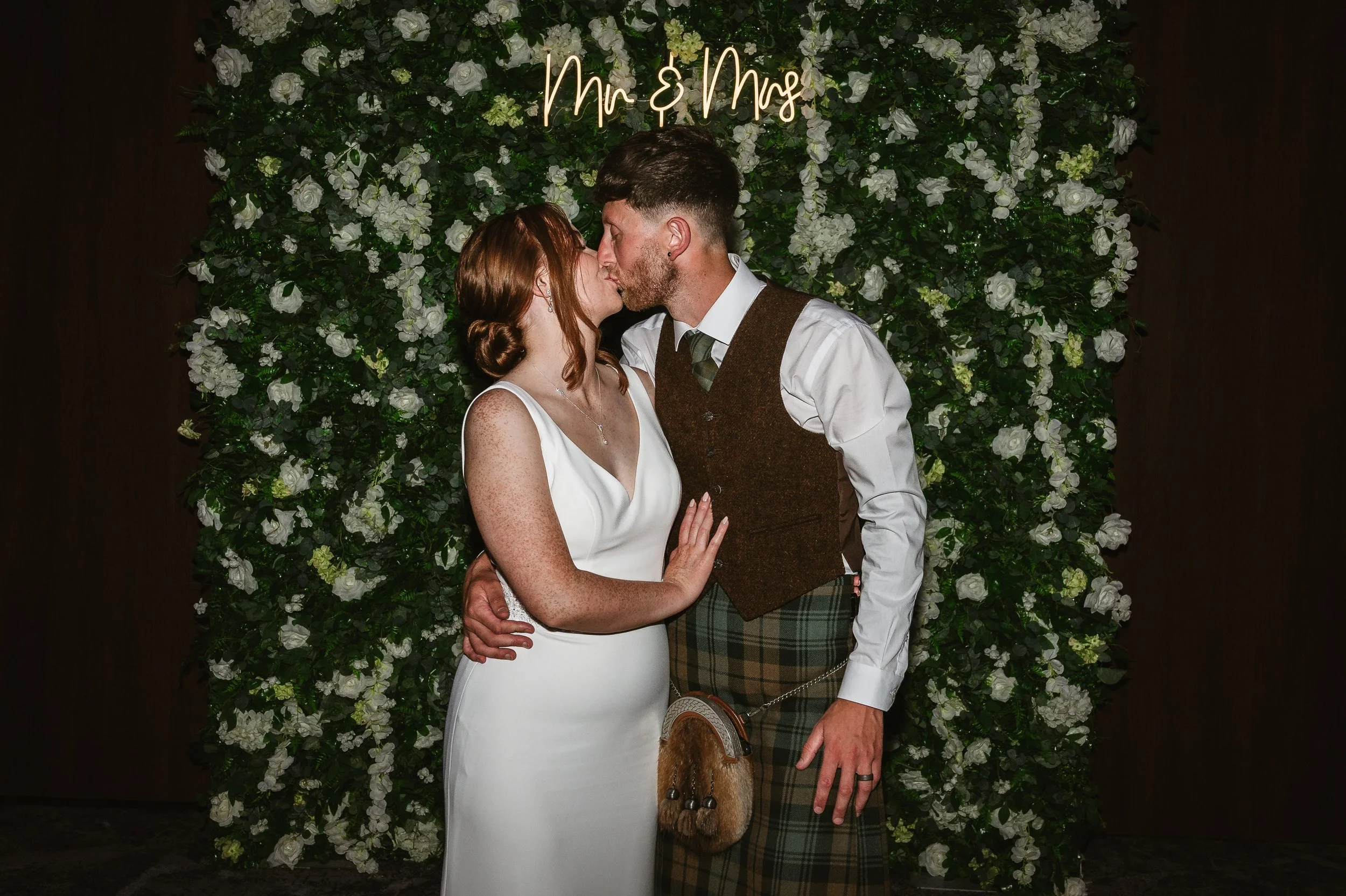 A couple kissing in front of a floral backdrop with a neon sign that reads 'Mr & Mrs'. The woman has red hair in a bun and is wearing a white wedding dress. The man has short dark hair, a beard, and is wearing traditional Scottish attire, including a