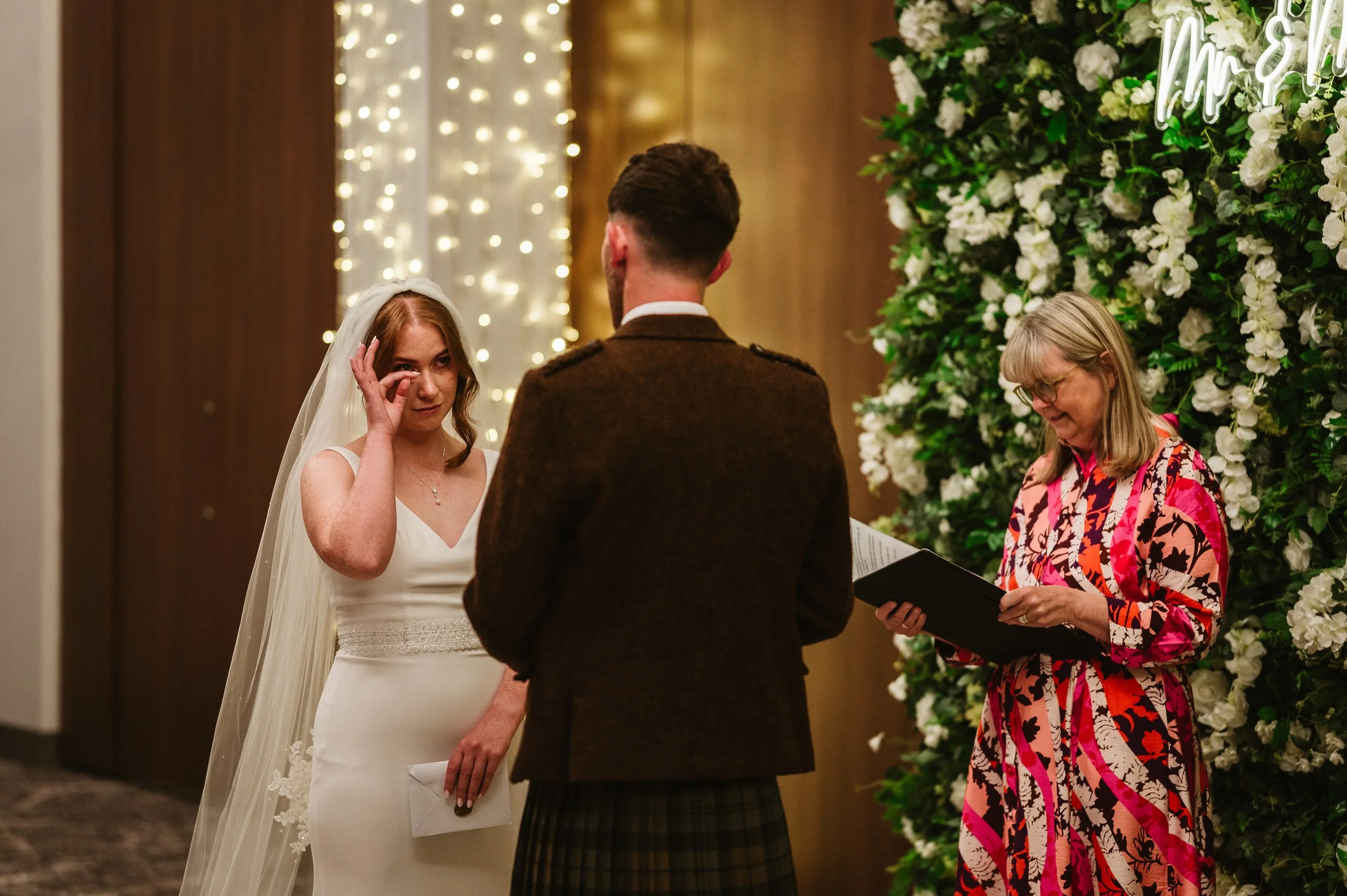 A bride wearing a white wedding dress and veil touching her eye during her wedding ceremony, with a man in a brown jacket and a woman in floral attire reading from a book beside a floral backdrop.