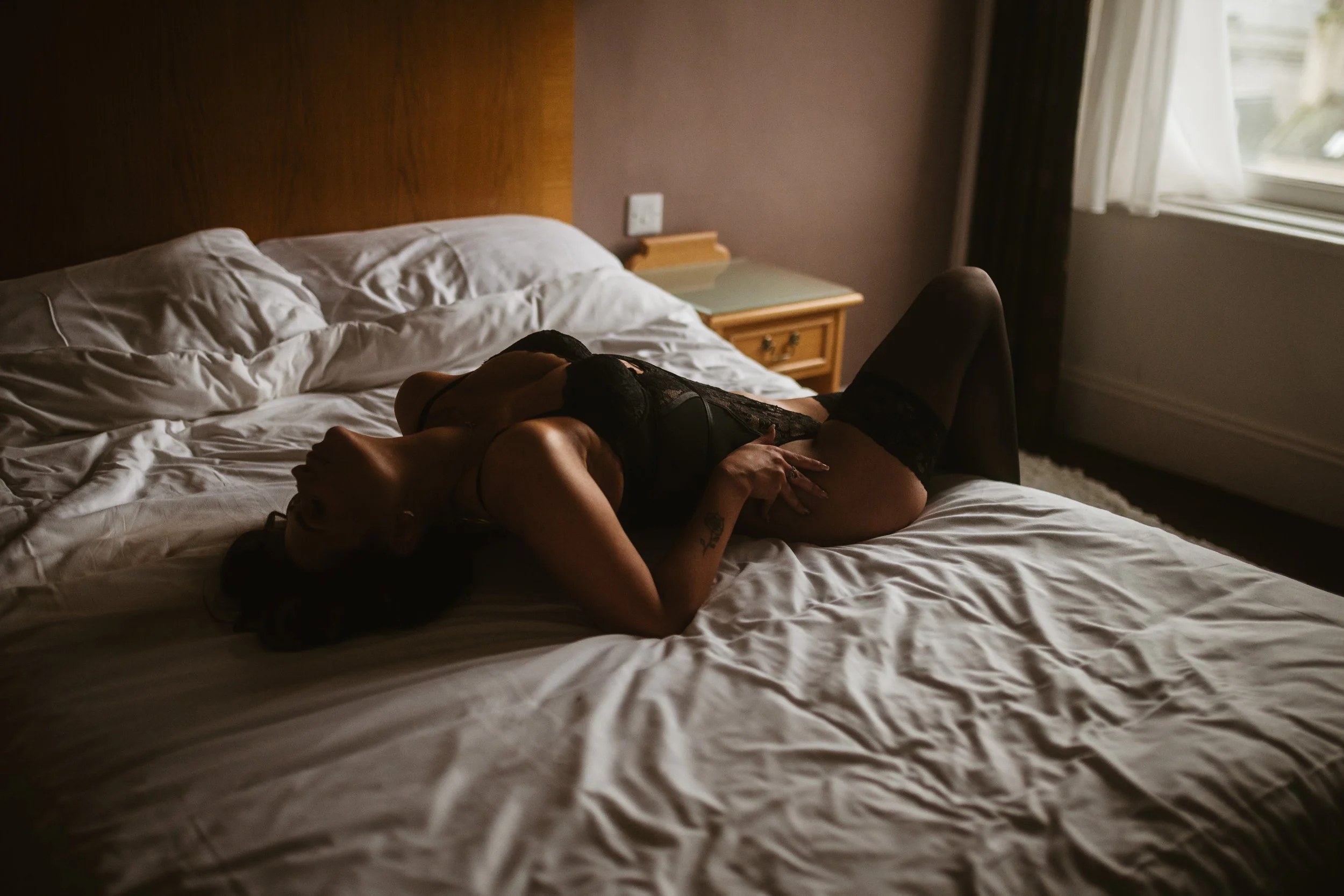A woman lying on a hotel bed in black lingerie, with natural light coming through a window.