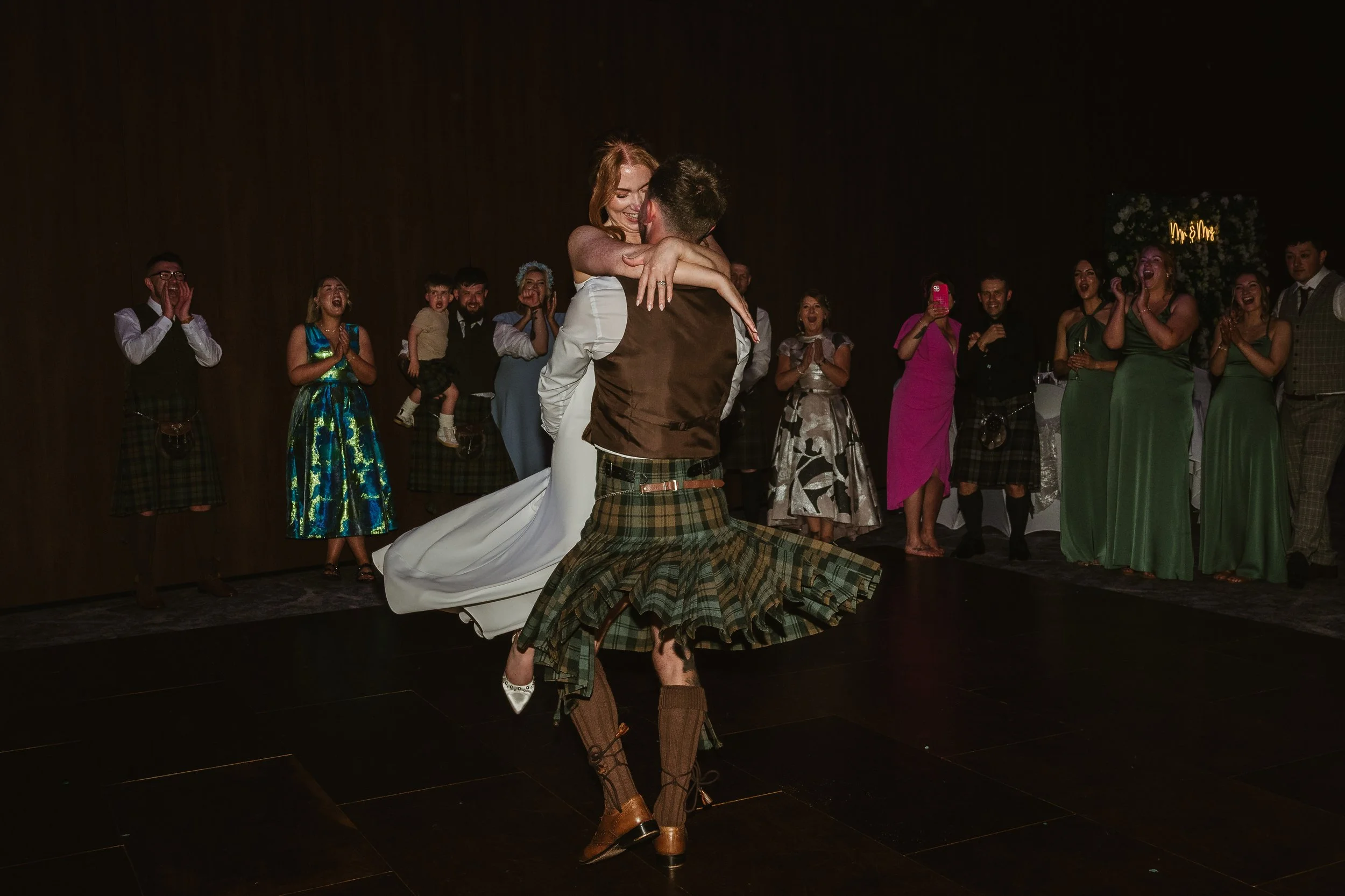 A bride and groom dancing closely at their wedding reception, surrounded by excited guests in formal attire, with some taking photos.