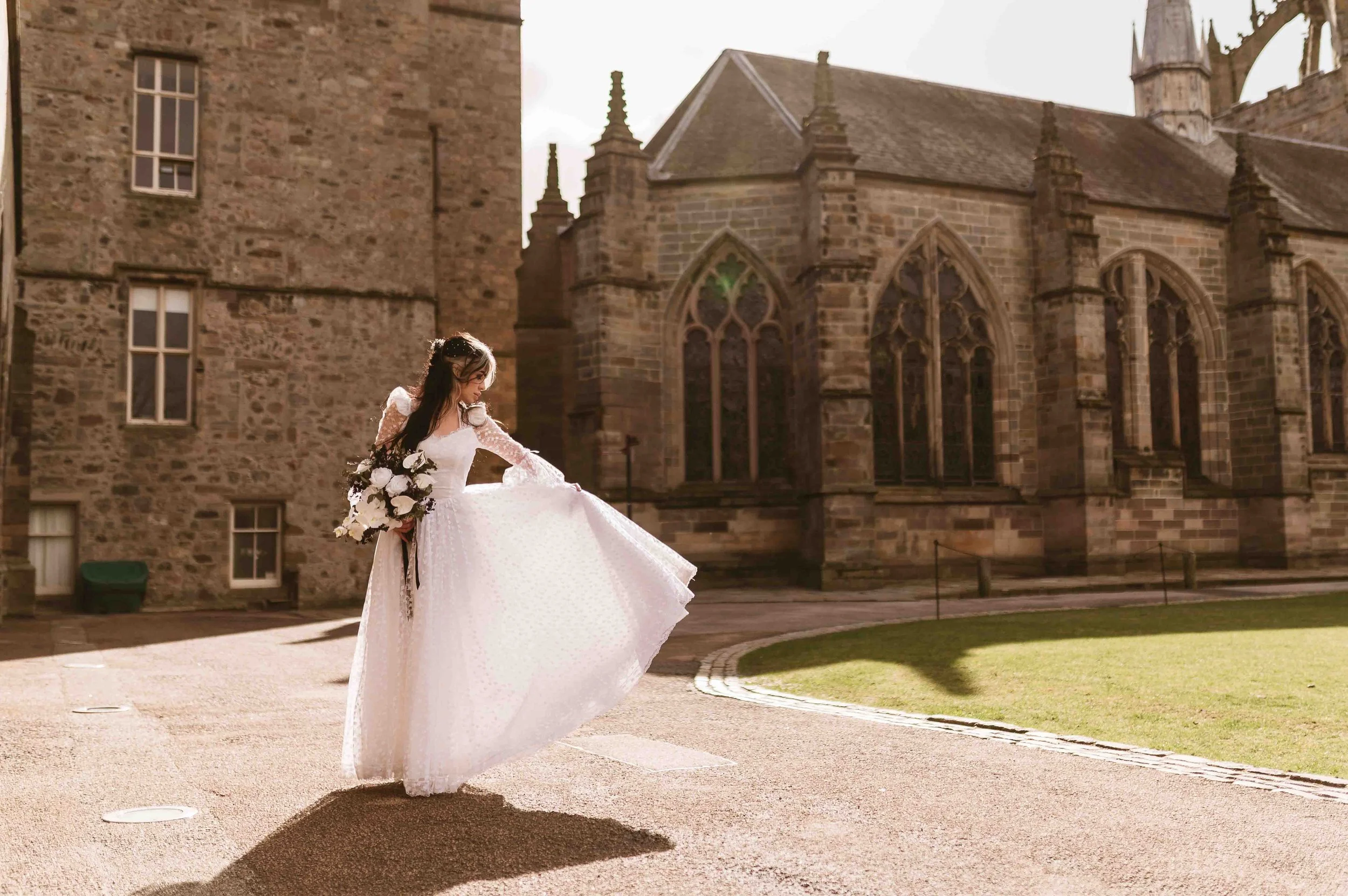 Bride in a white wedding dress holding a bouquet, standing outdoors in front of a historic stone church or building in sunlight.