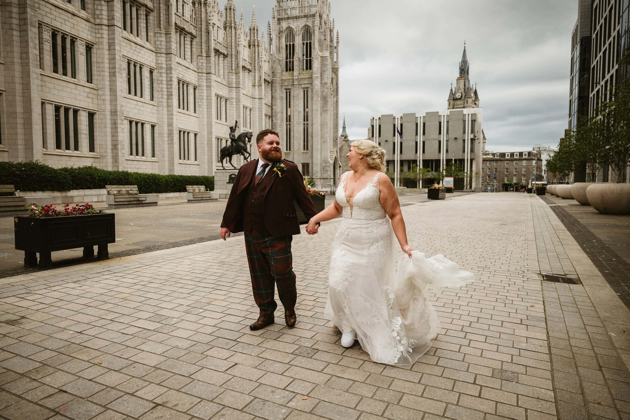 A newly married couple holding hands and smiling while walking outdoors in front of a historic cathedral with gothic architecture in a city square, under an overcast sky.