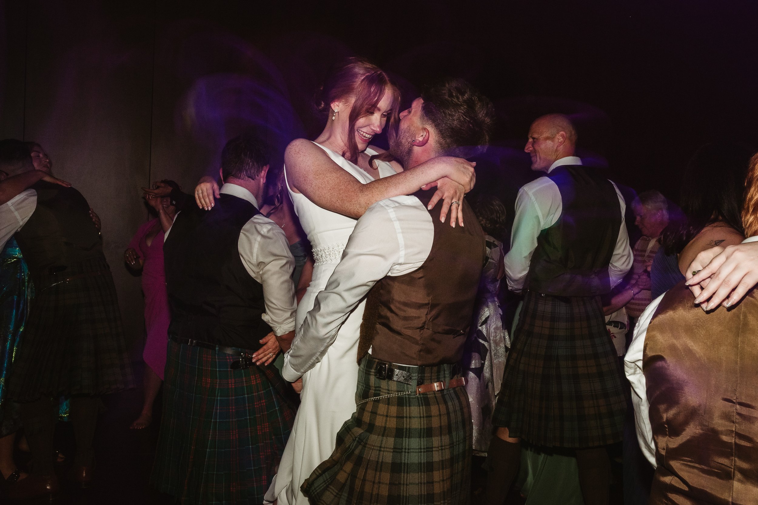 A couple dancing closely and smiling at an indoor celebration, surrounded by other people in formal attire, with dim lighting and purple hues.