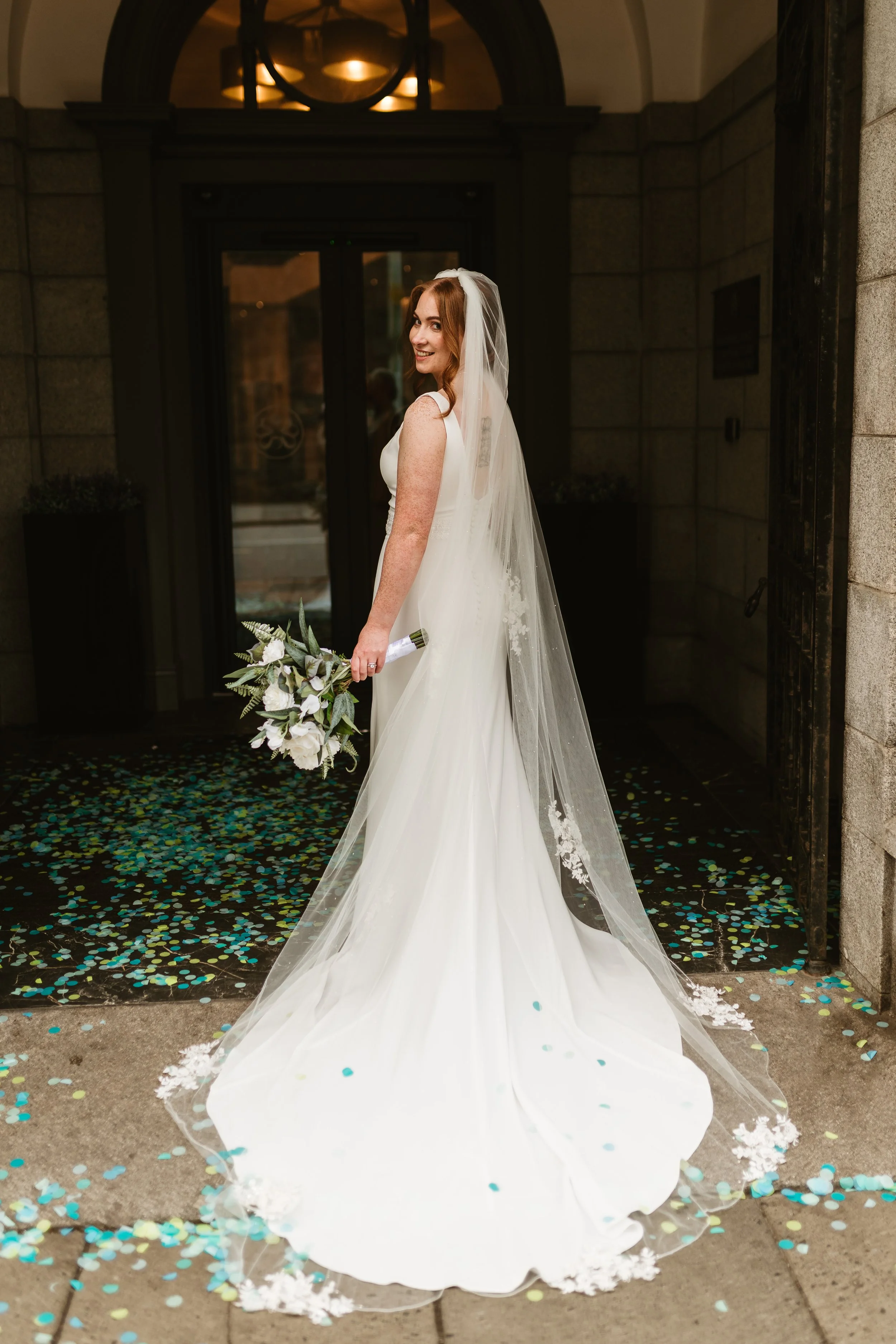 A bride standing outdoors in front of a building doorway holding a bouquet of white flowers and greenery, wearing a white wedding gown and veil, with confetti scattered on the ground.