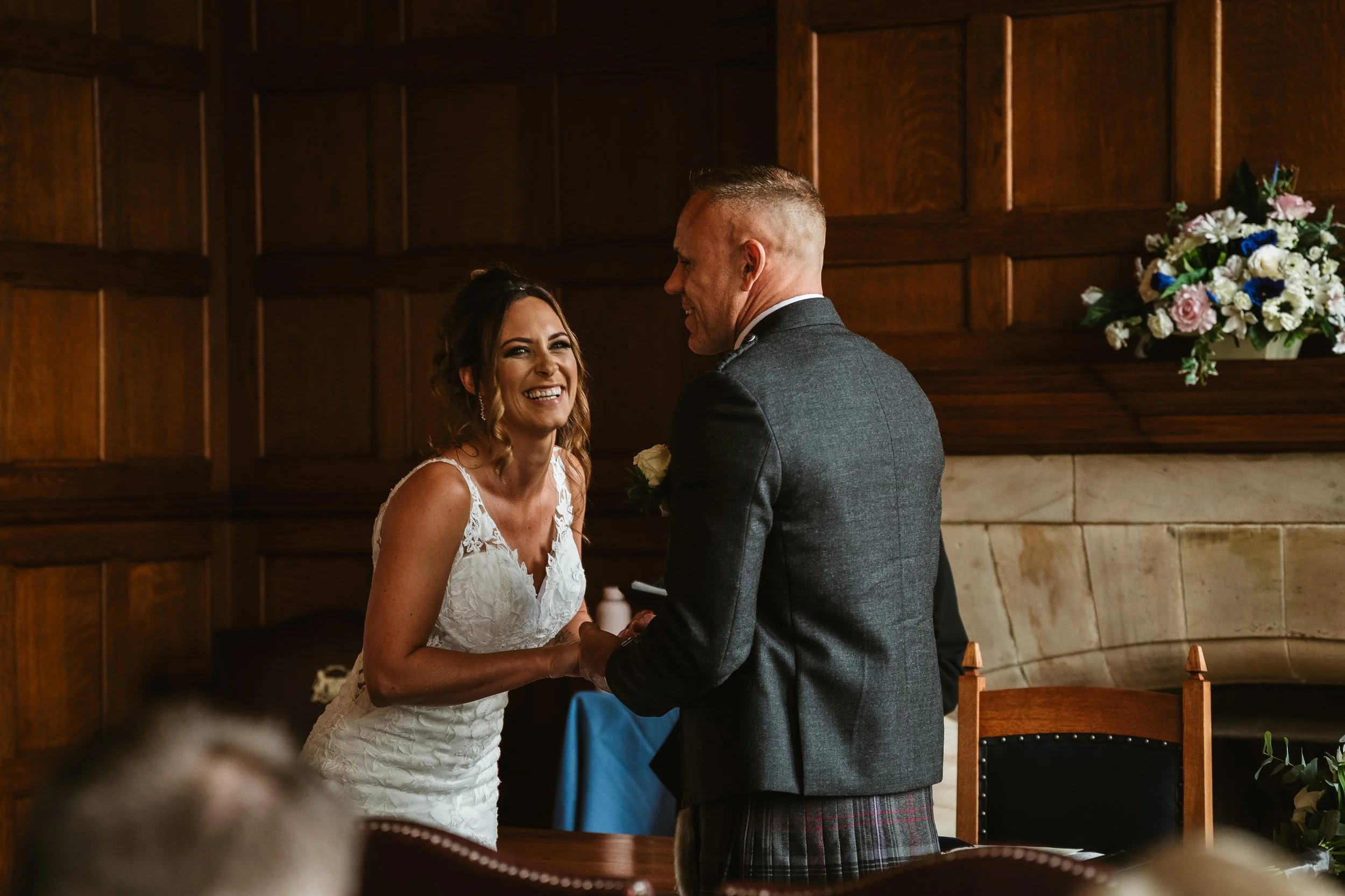 A bride and groom holding hands during their wedding ceremony, smiling at each other in a wood-paneled room with a floral arrangement in the background.