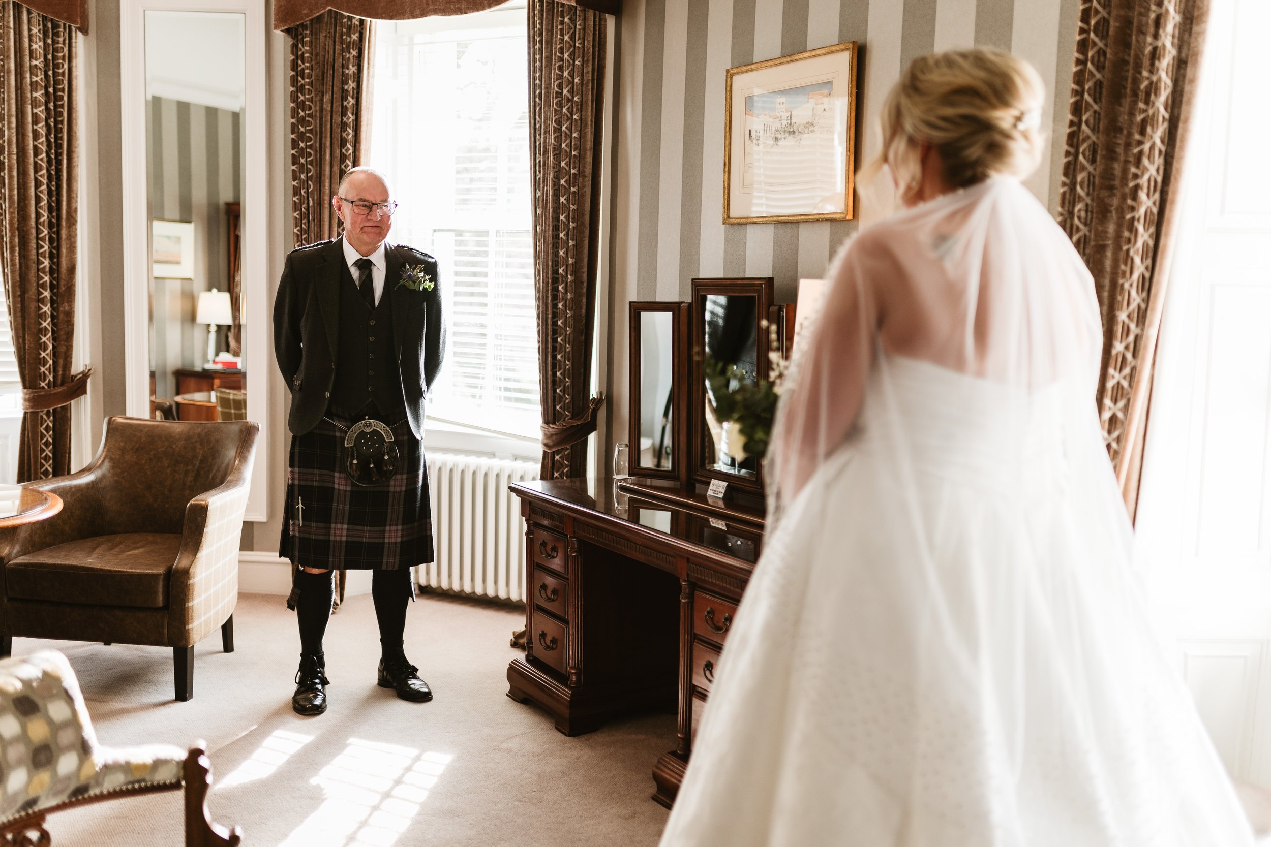 A bride and an older man in a room with large windows and vintage furniture, some mirrors on a dresser, and framed pictures on the wall. The man is dressed in traditional Scottish attire with a kilt, black jacket, and glasses, standing with his hands