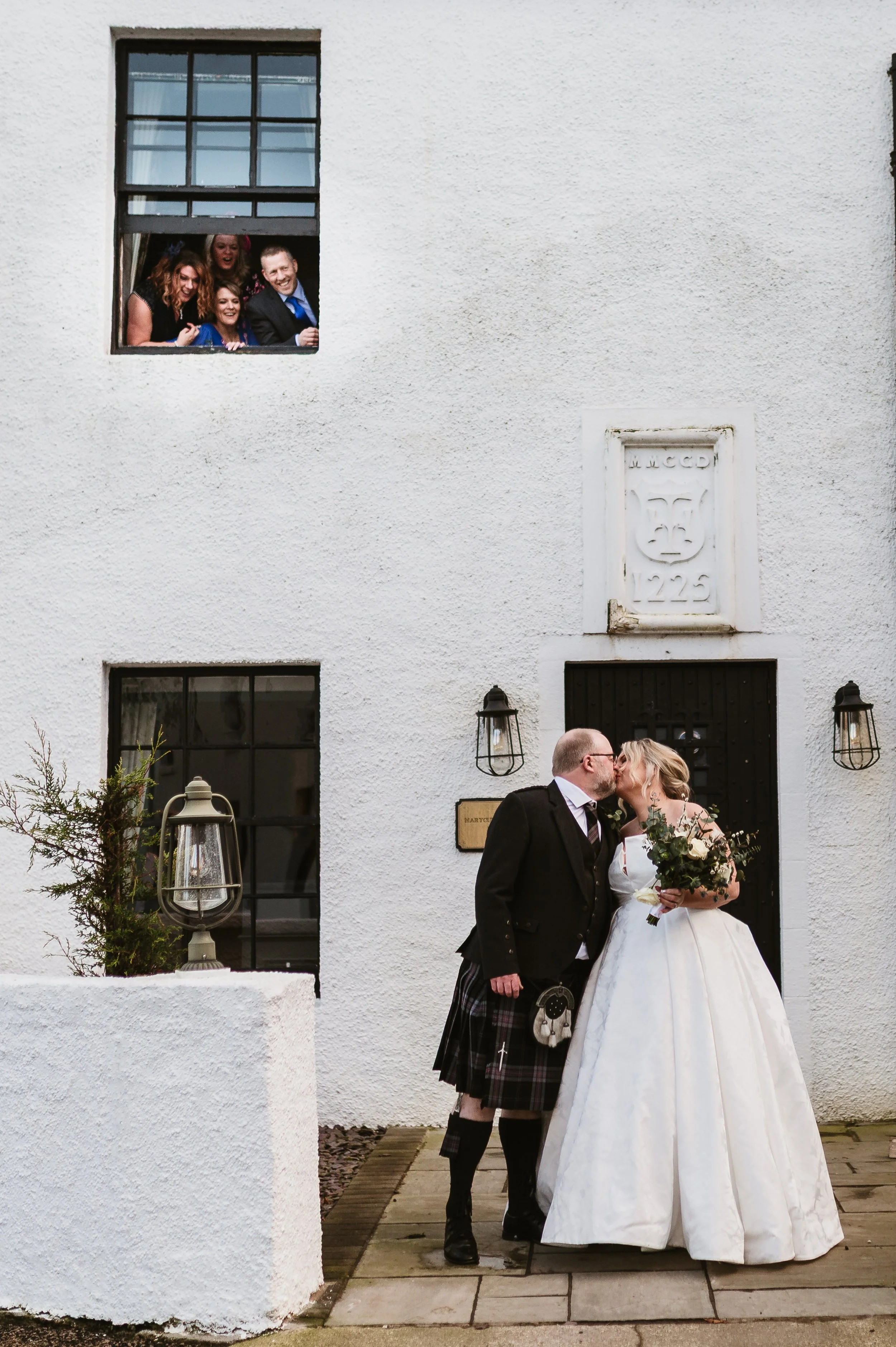 A wedding couple sharing a kiss in front of a white building. The bride is holding a bouquet and wearing a white wedding dress, and the groom is dressed in a kilt and tuxedo. There are two black lantern lights mounted on the wall and a small plant in