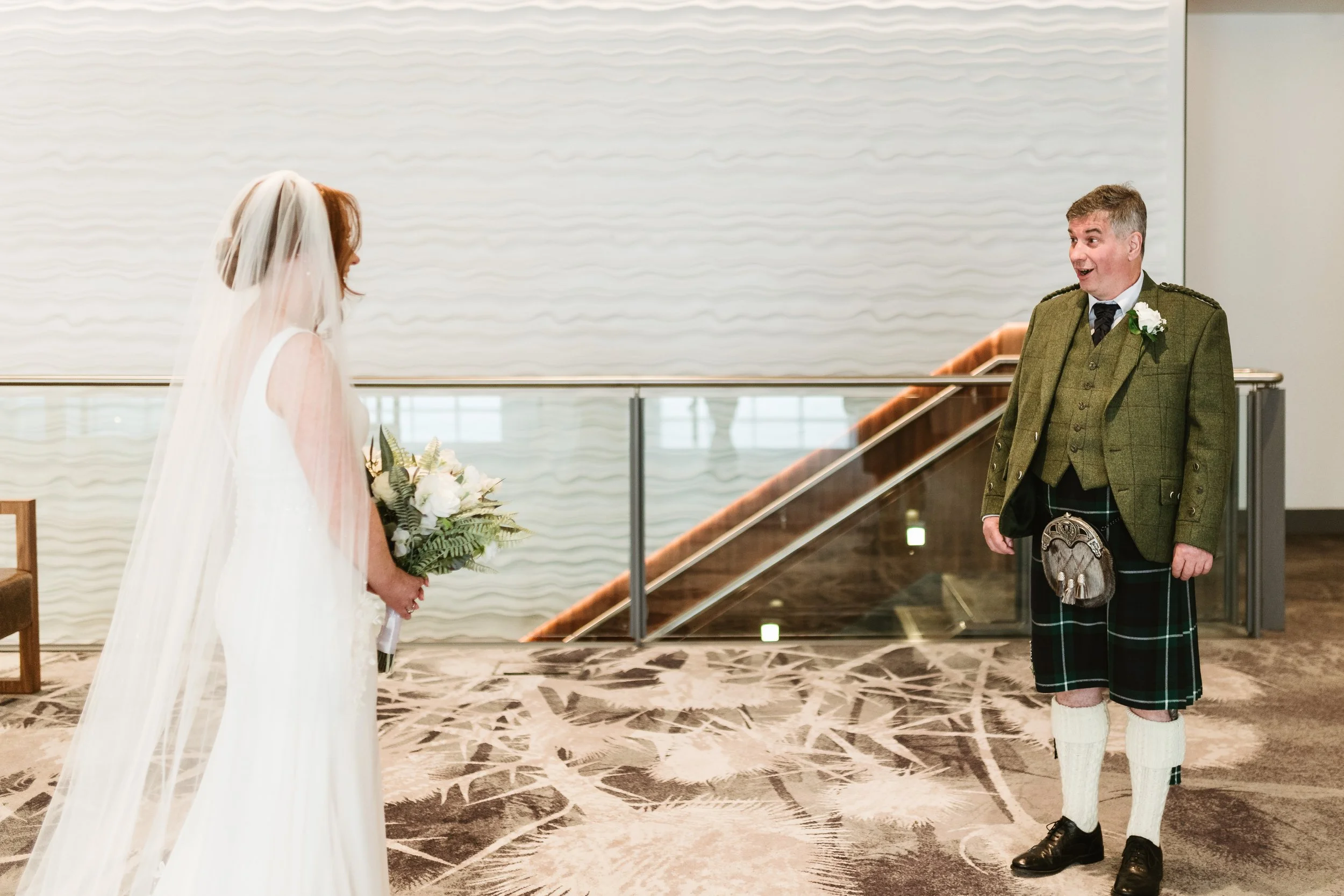 A bride in a white wedding gown with a veil holding a bouquet of white flowers, standing and facing a man dressed in traditional Scottish attire, who is smiling and looking at her, inside a modern venue with a staircase in the background.