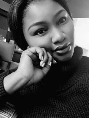 A young woman Dirichi O Phillips with short hair, wearing a dark sweater, resting her chin on her hand, looking at the camera.