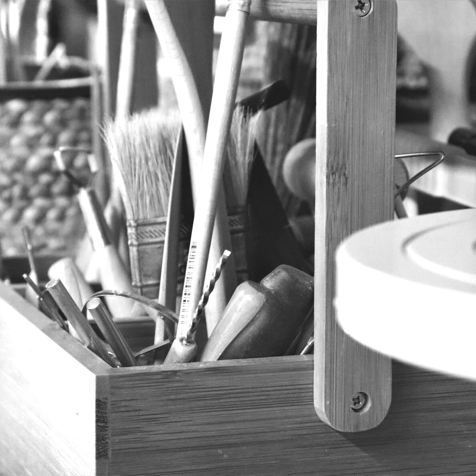 A wooden toolbox filled with various tools, including paintbrushes, screwdrivers, and a chisel, placed on a workbench with wood shelves in the background.