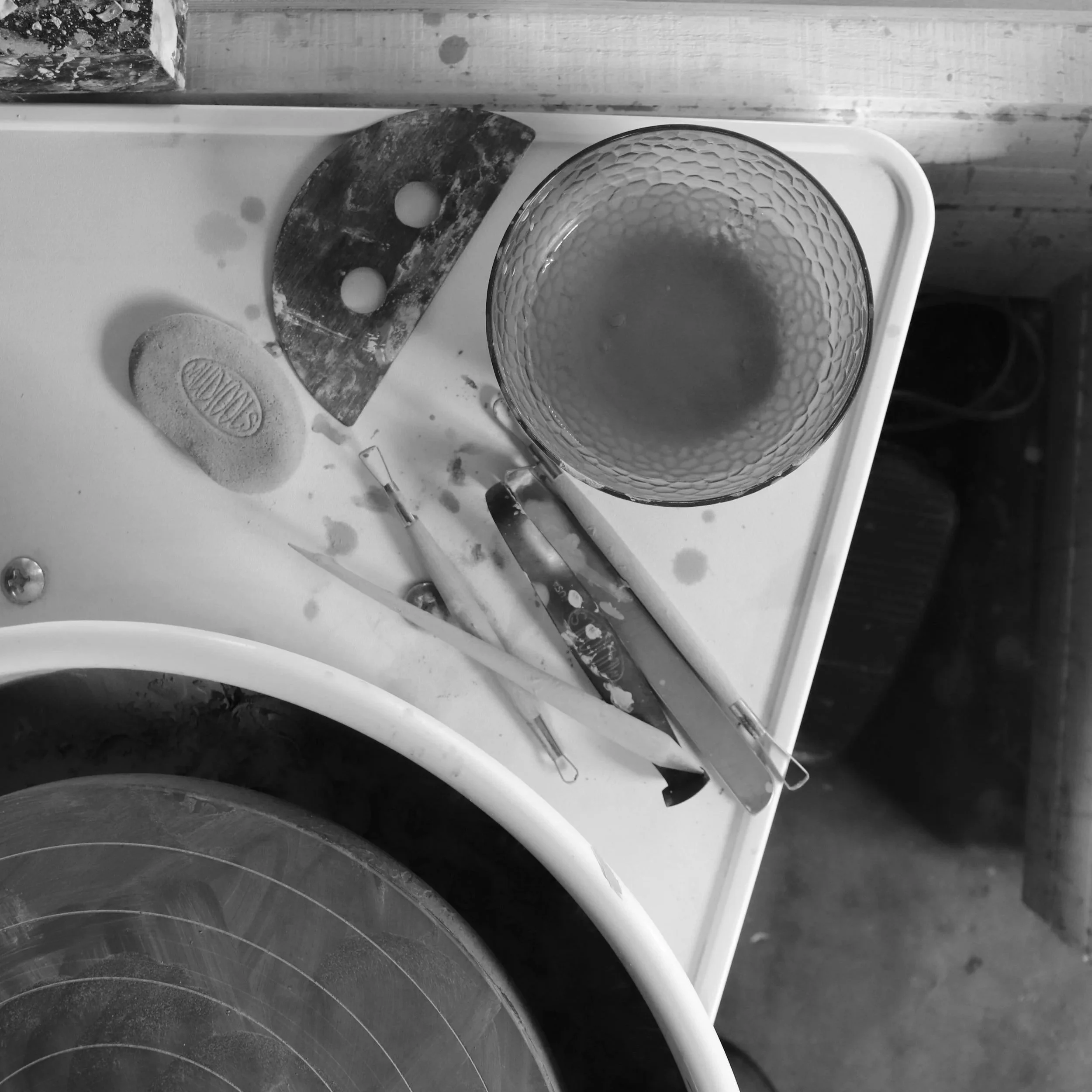 Top-down view of a cluttered work surface with a glass bowl, a sculpting tool, and a spatula, surrounded by other tools and materials, likely for artistic or kitchen use.