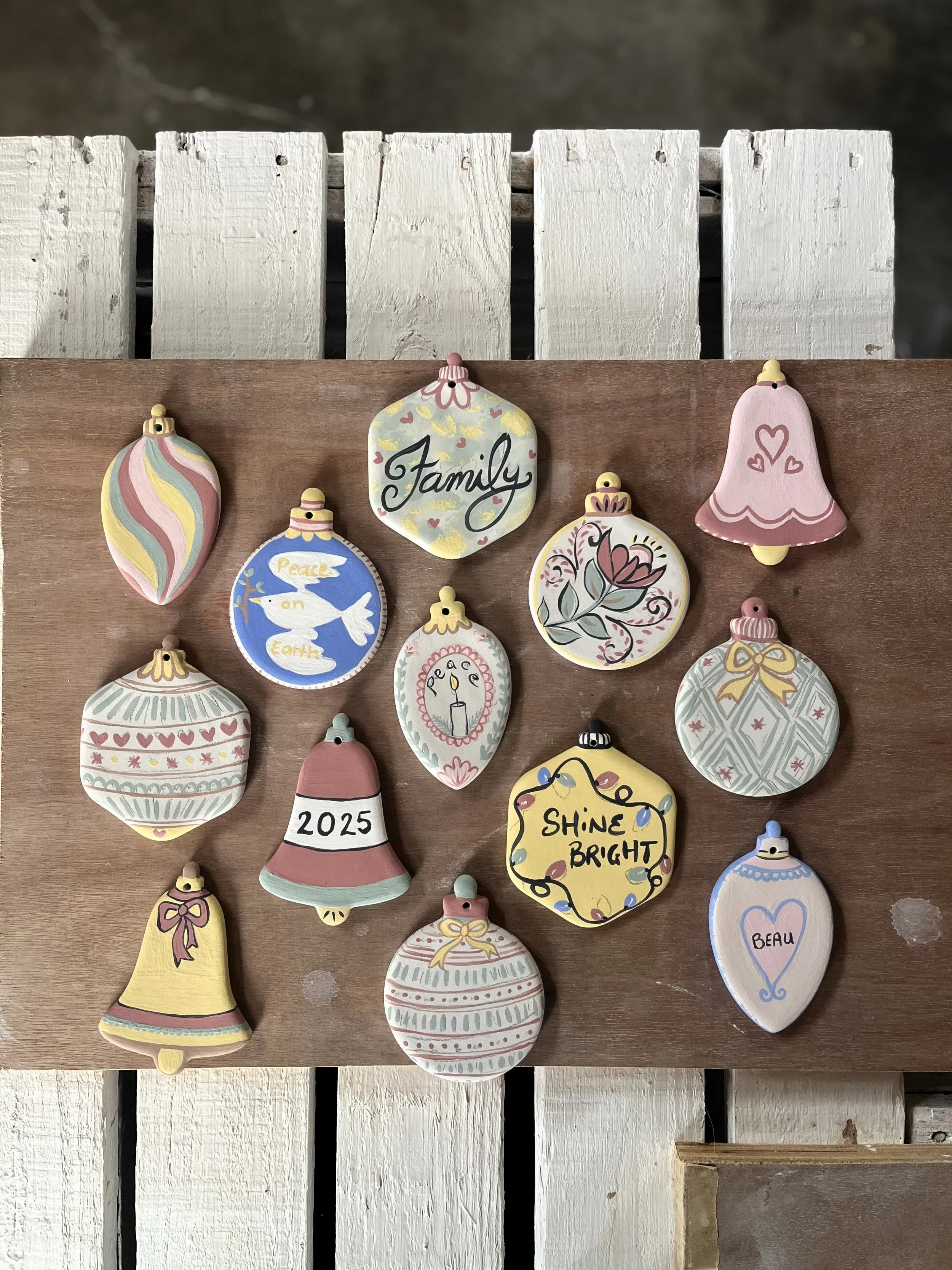 Decorative holiday Christmas ornaments on a wooden surface, including a 'Family' ornament, a dove with 'Peace on Earth' message, a candle, and a 'Shine Bright' ornament, among others for a hand painting ceramic class.