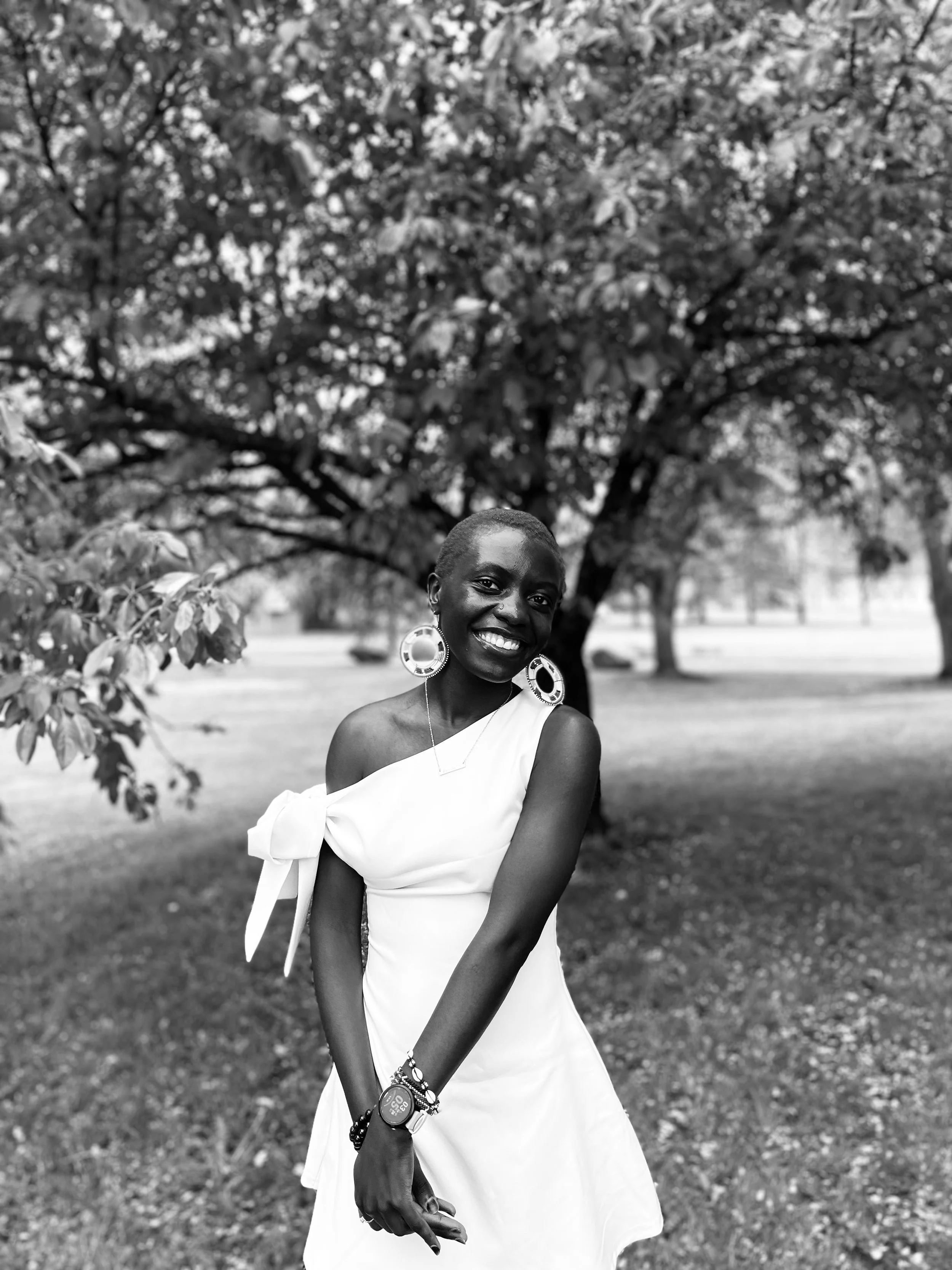 A smiling woman in a white dress with large earrings standing outdoors in front of a large tree in a park.