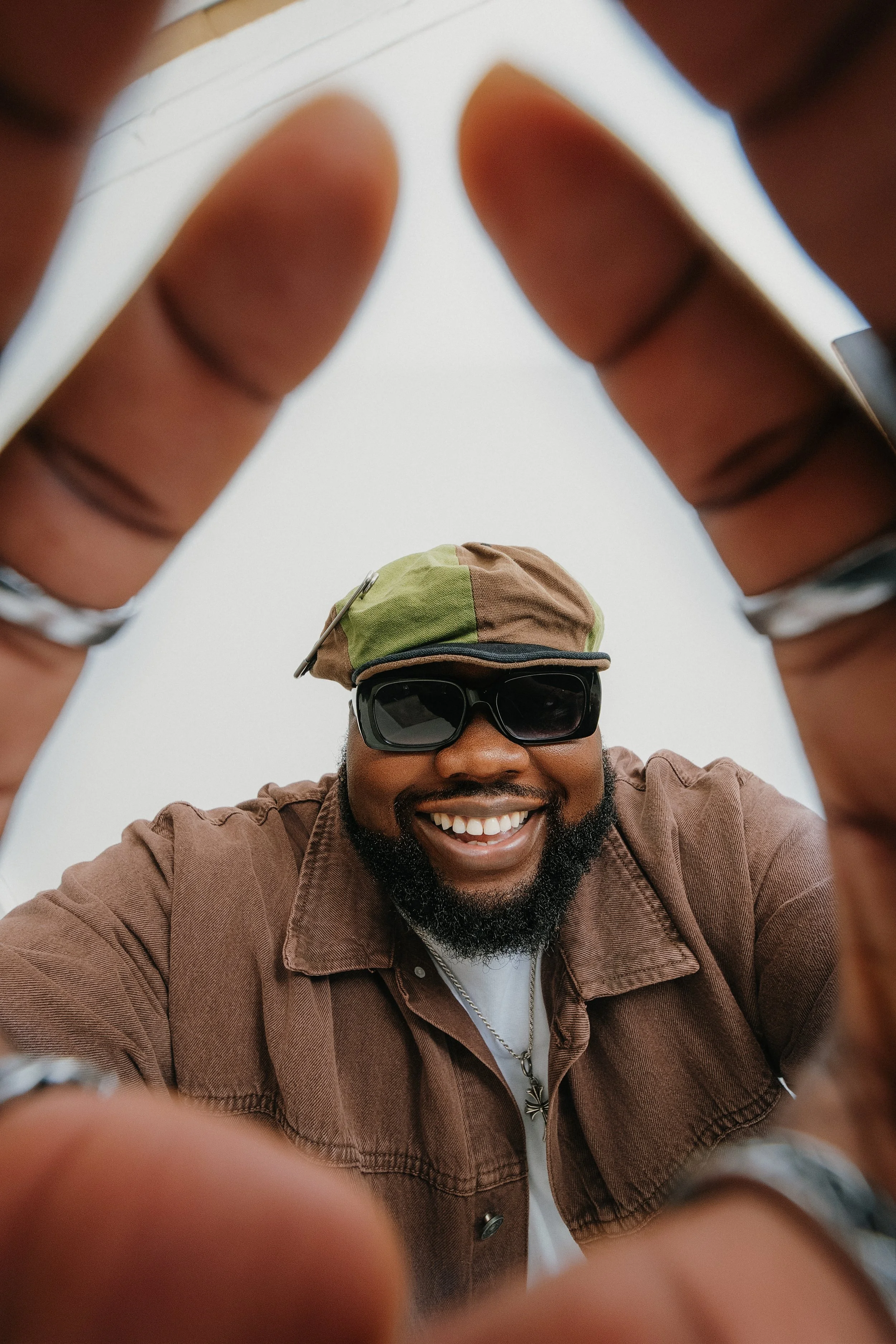 A man smiling, wearing sunglasses and a patterned cap, is seen from below through the frame of his fingers, with a plain sky in the background.