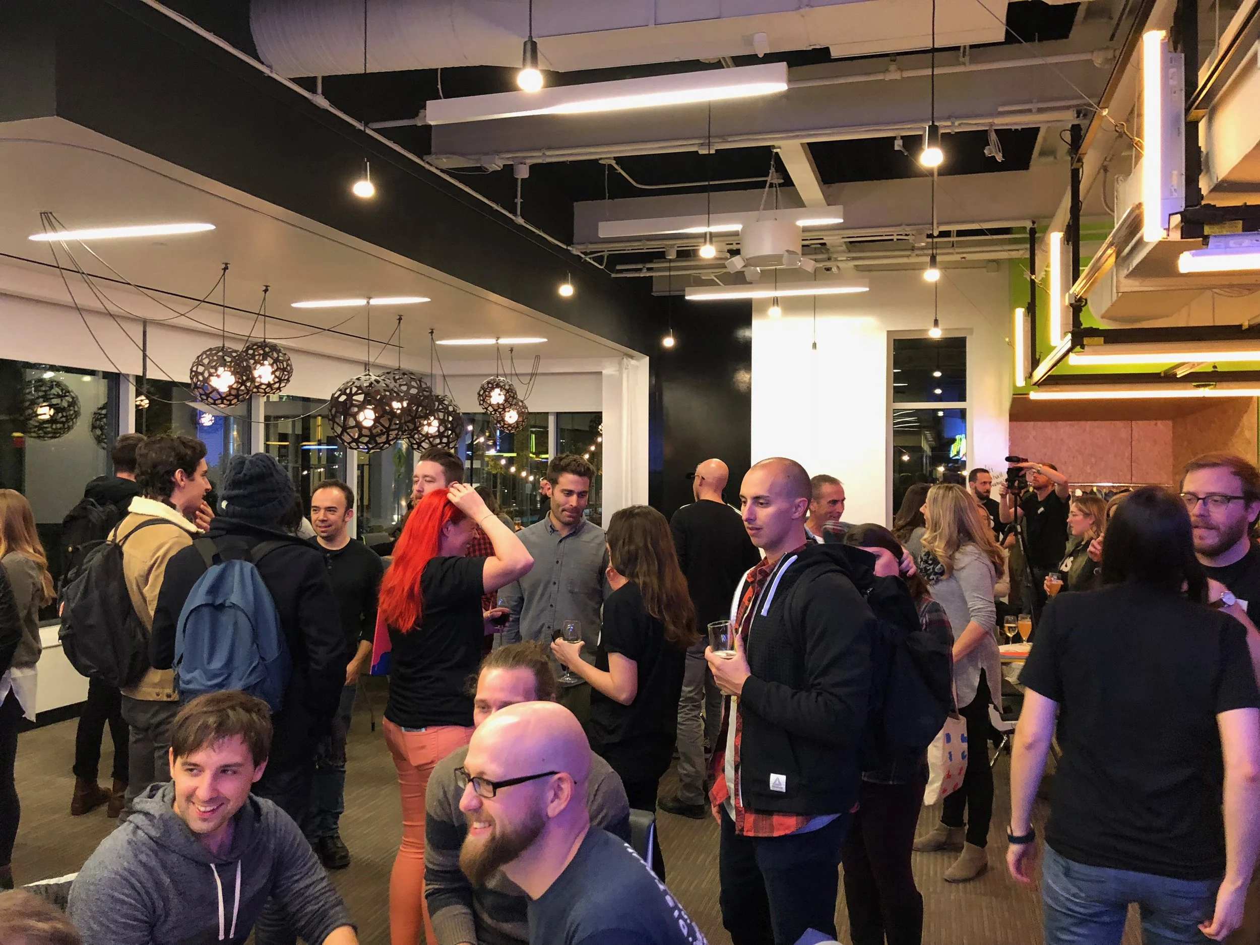 People socializing at a casual indoor event or party, with some holding drinks and engaging in conversation, in a room decorated with modern pendant lights.