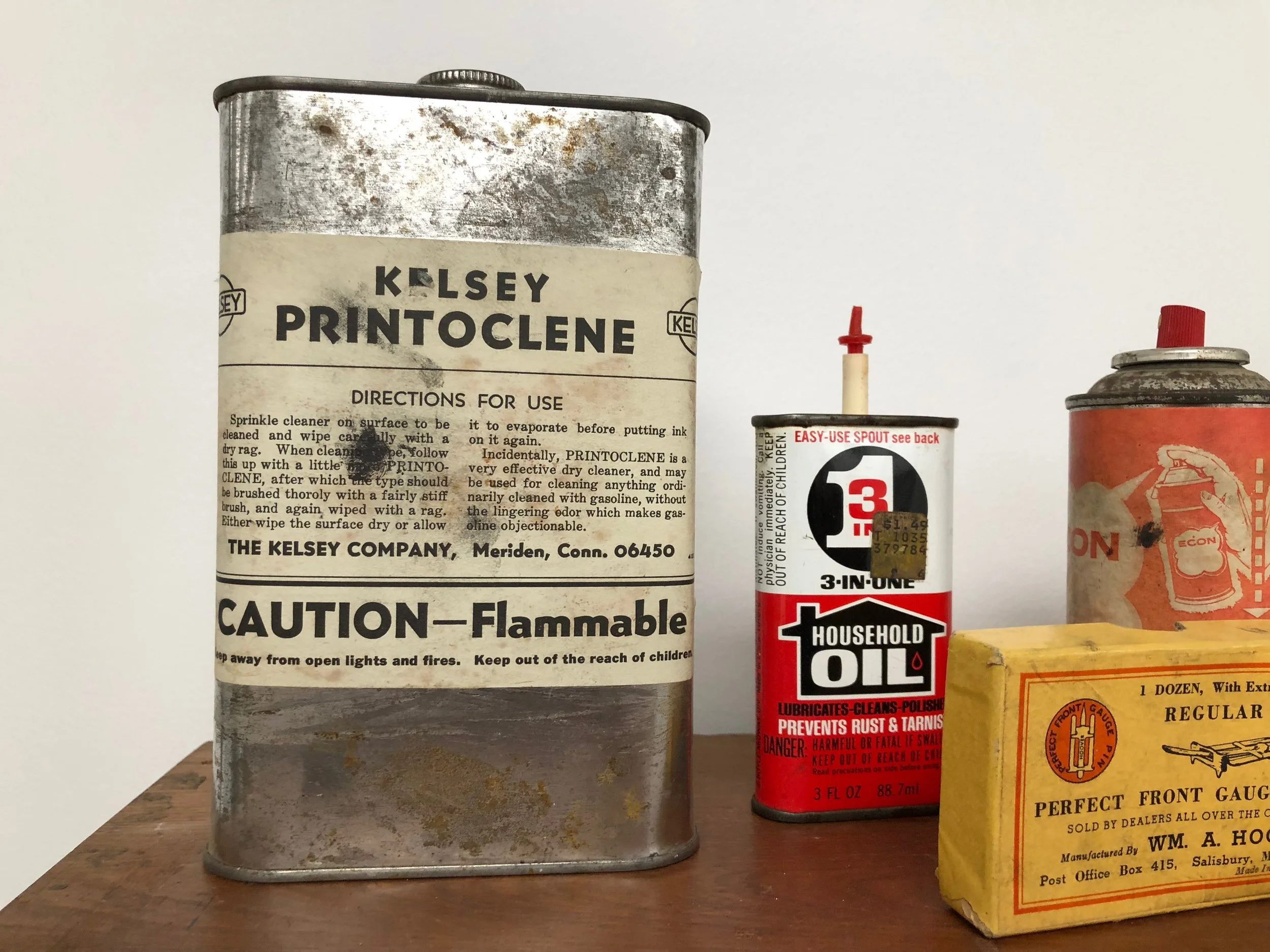 A collection of vintage household items on a wooden surface, including a large metal can of Kelsey Printoclene, a small red and white spray can of Household Oil, a cylindrical can with a red cap, and a yellow box labeled Wm. A. Hodge.