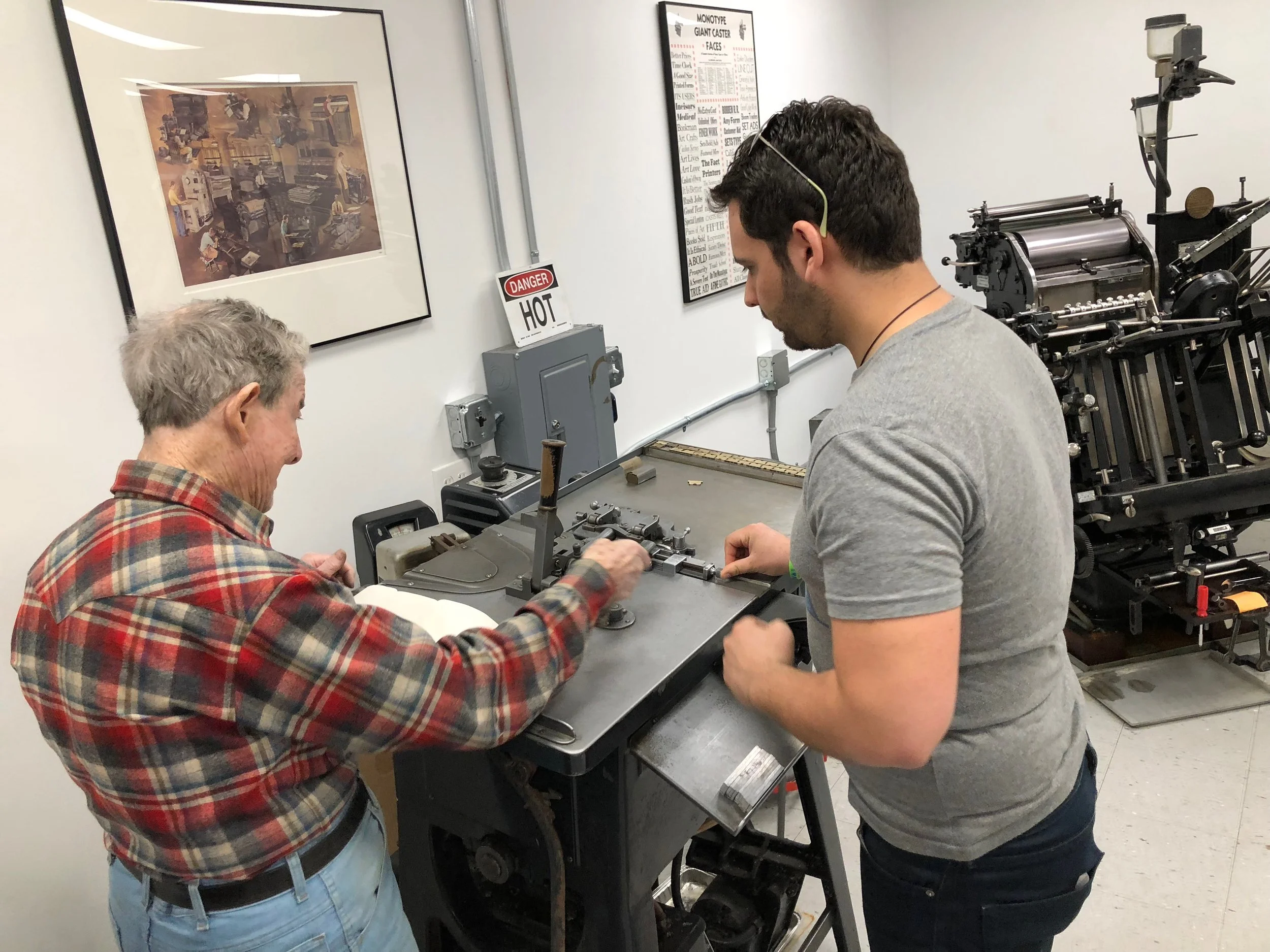 Two men working at a printing press or similar machine in a workshop or studio, with posters on the wall and safety signs.