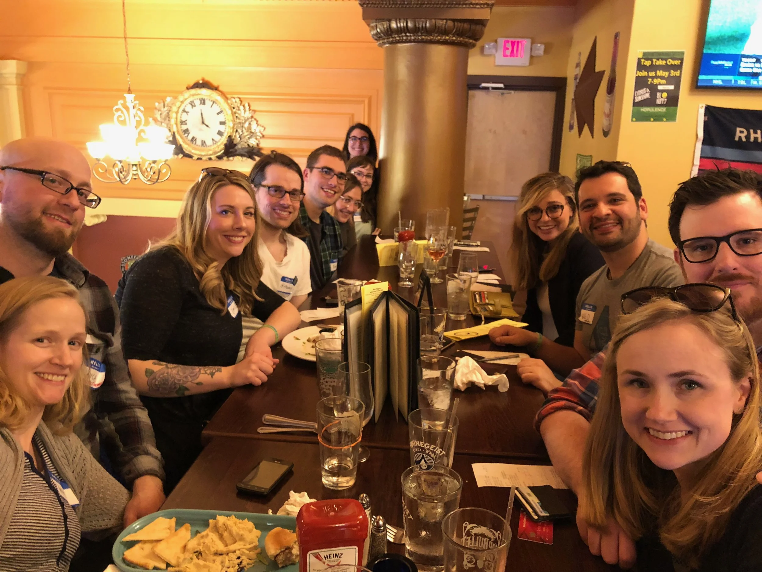 Group of people sitting at a long restaurant table, smiling for a group photo, with plates, glasses, and condiments visible on the table, illuminated by warm lighting with decorative wall elements in the background.