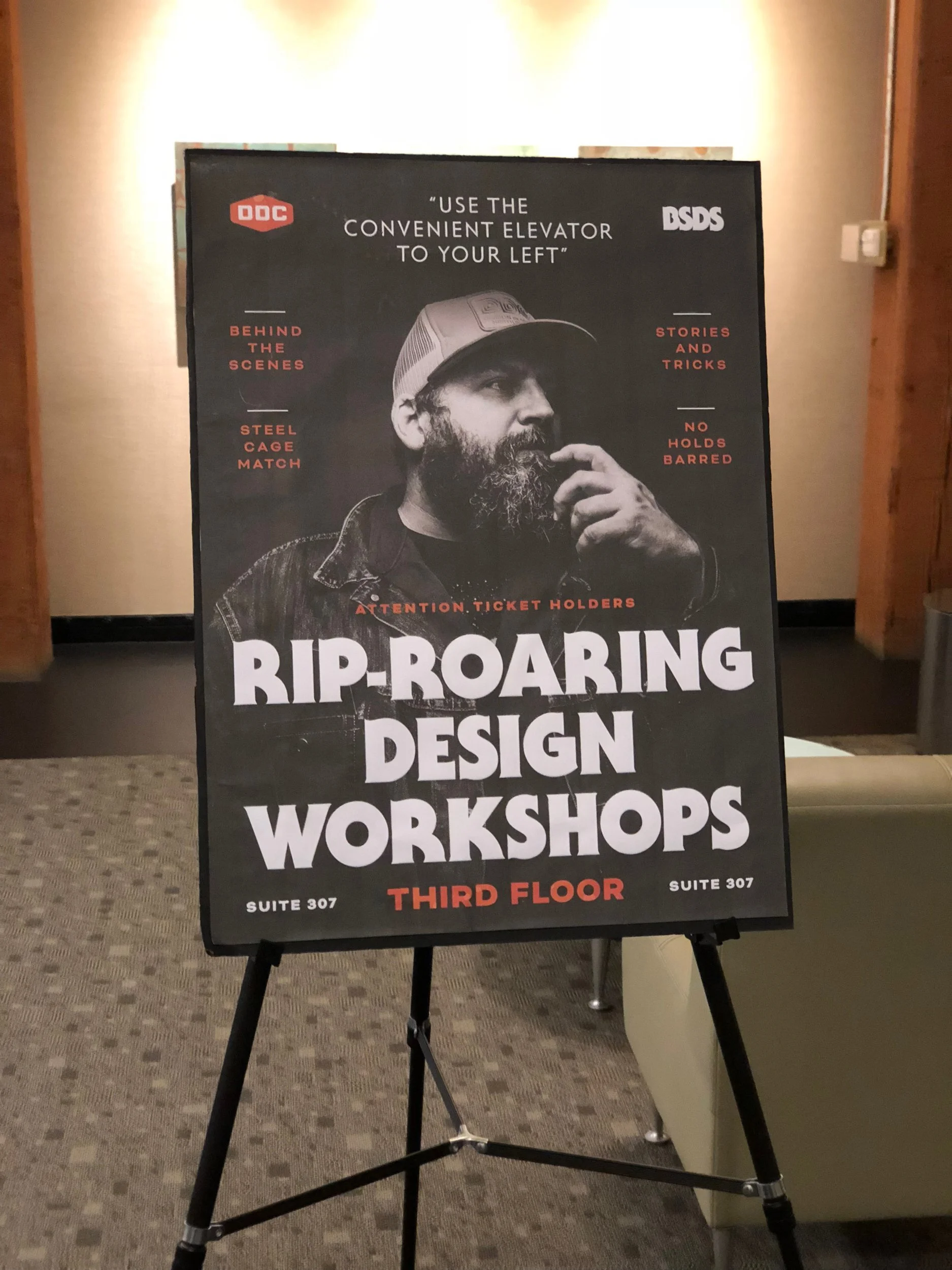 A black sign promoting rip-roaring design workshops on the third floor, Suite 307, with an image of a man with a beard and baseball cap and phrases about elevator use and workshop details.