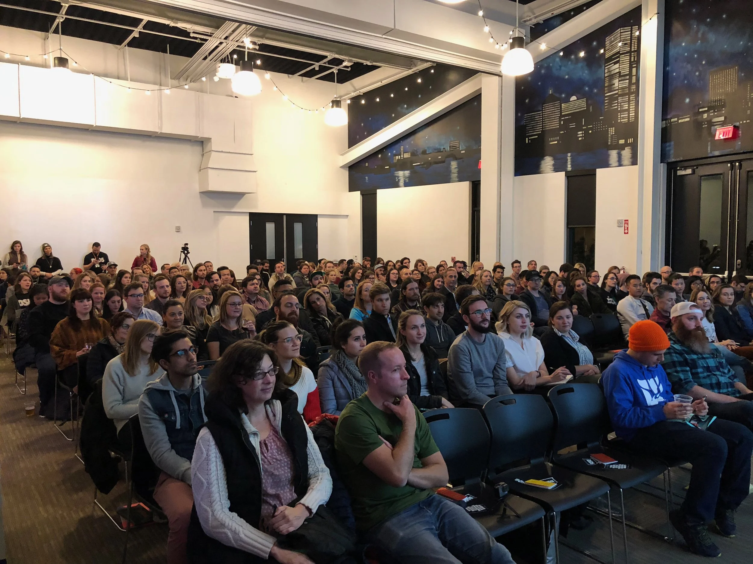 Audience attending a presentation or event in a large indoor venue with wall art of a city skyline at night.