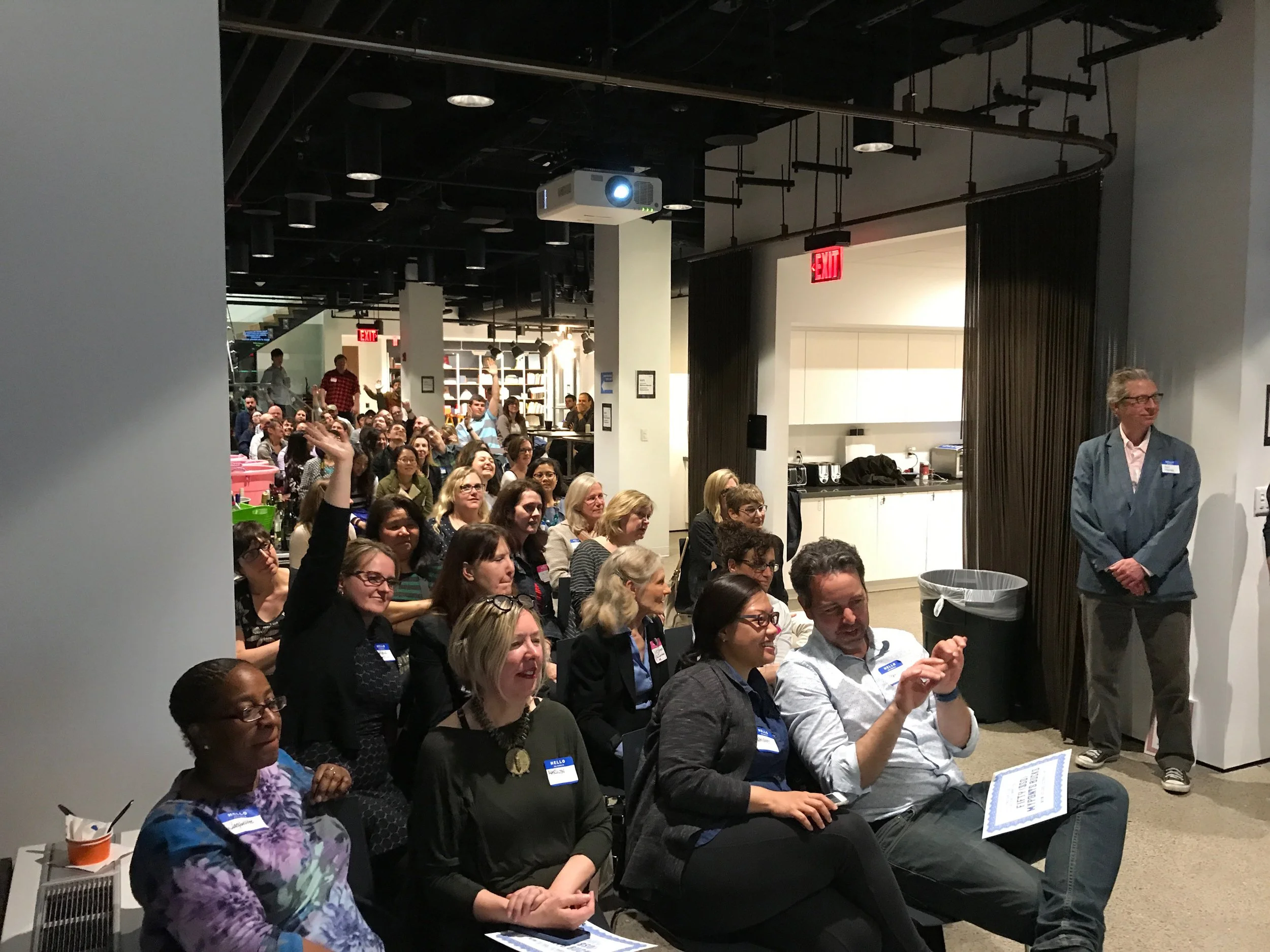 An indoor event with a seated audience of diverse adults attentively listening, some smiling and one woman raising her hand, in a room with a kitchenette and a projector overhead.