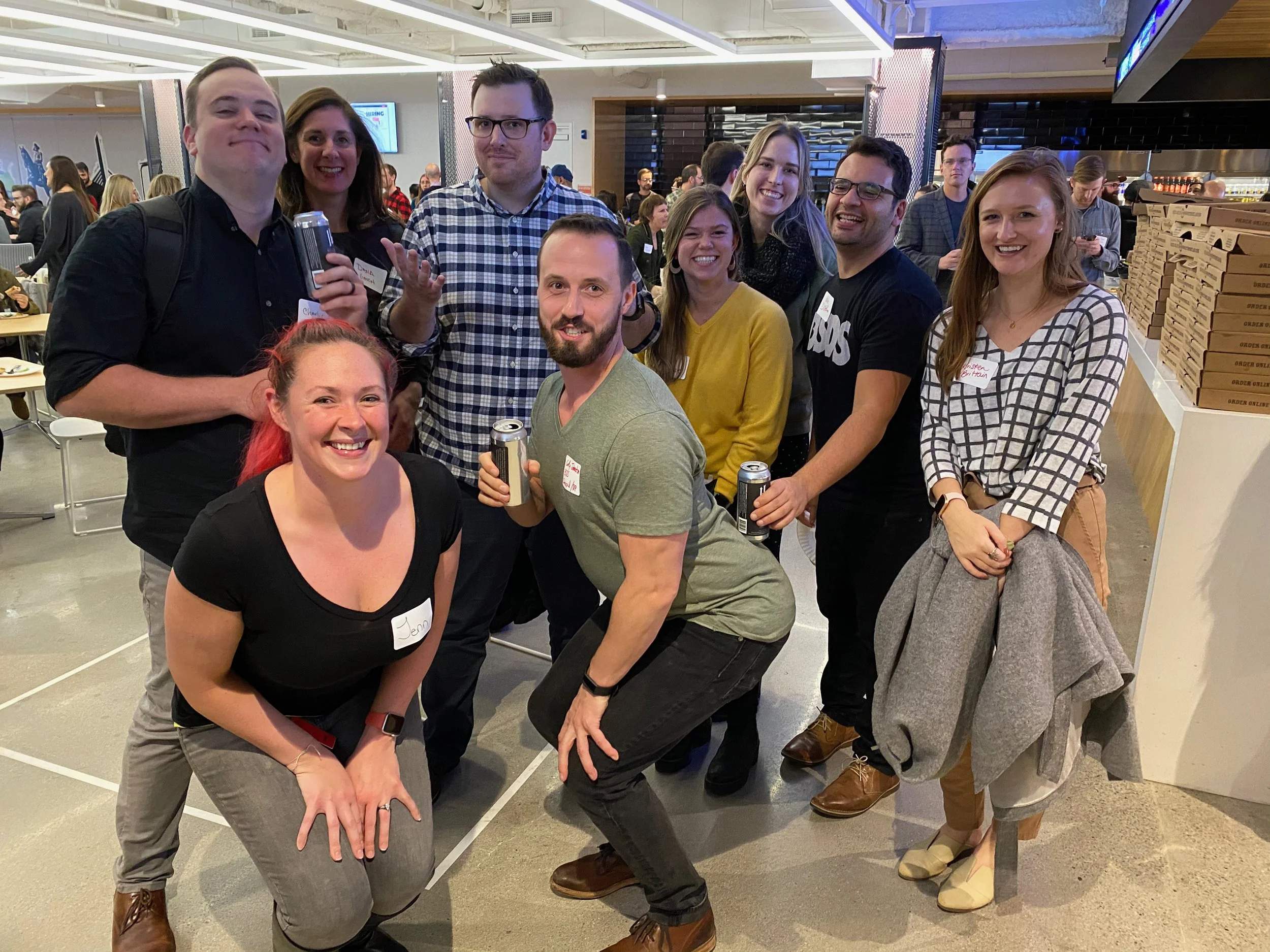 Group of ten people smiling and posing for a photo at a social gathering or event, some holding drinks, in what appears to be a restaurant or event space with other people in the background.
