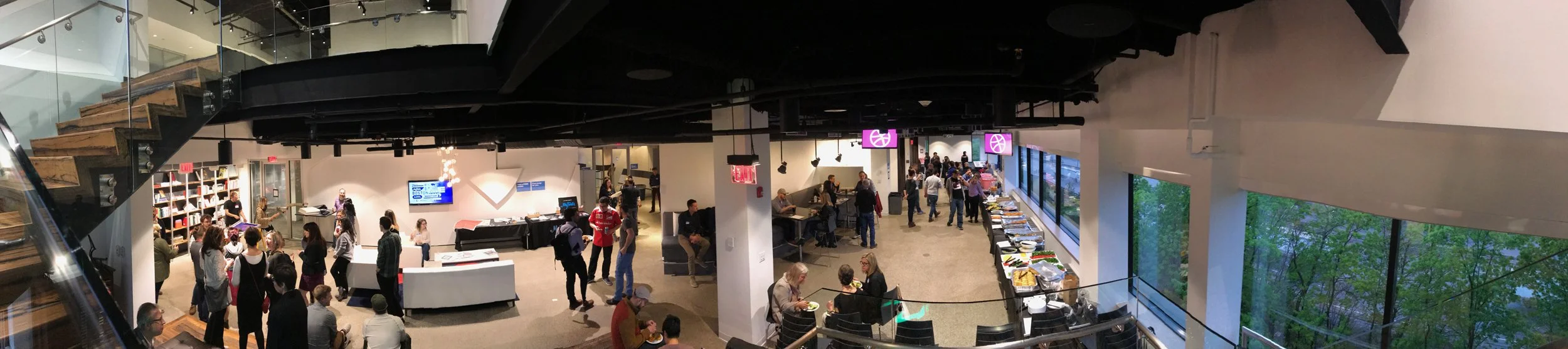 Interior of a busy modern cafeteria or lounge area with people sitting and walking around, large windows showing trees outside, and a staircase on the left side.