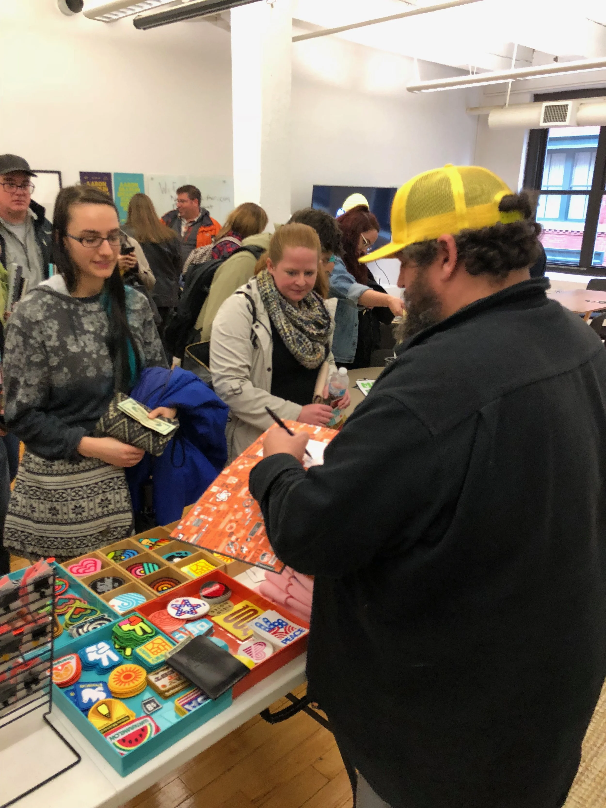 People standing in line at a table with colorful pins and trinkets, inside a room with white walls and large windows.