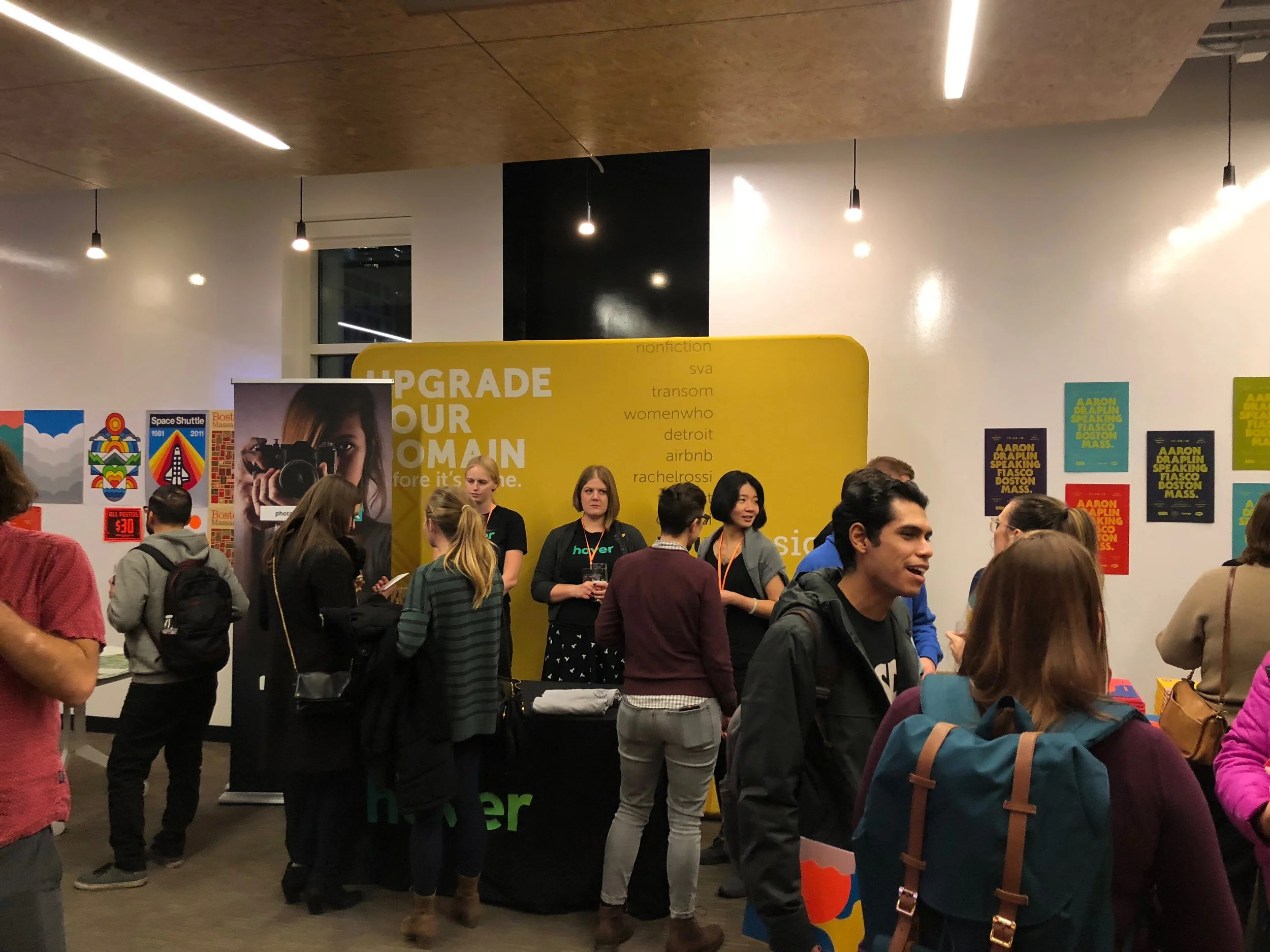 A group of people at a busy event booth with a yellow backdrop that has the text 'Upgrade your domain' and a list of popular domain extensions. Some people are engaging with the booth staff, while others are walking or talking.