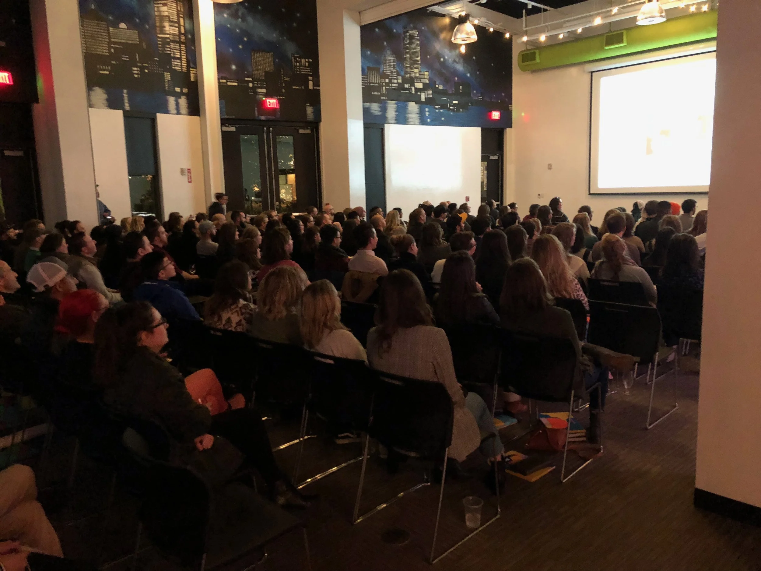 Large audience watching a presentation or movie in a dark room with a large screen at the front.