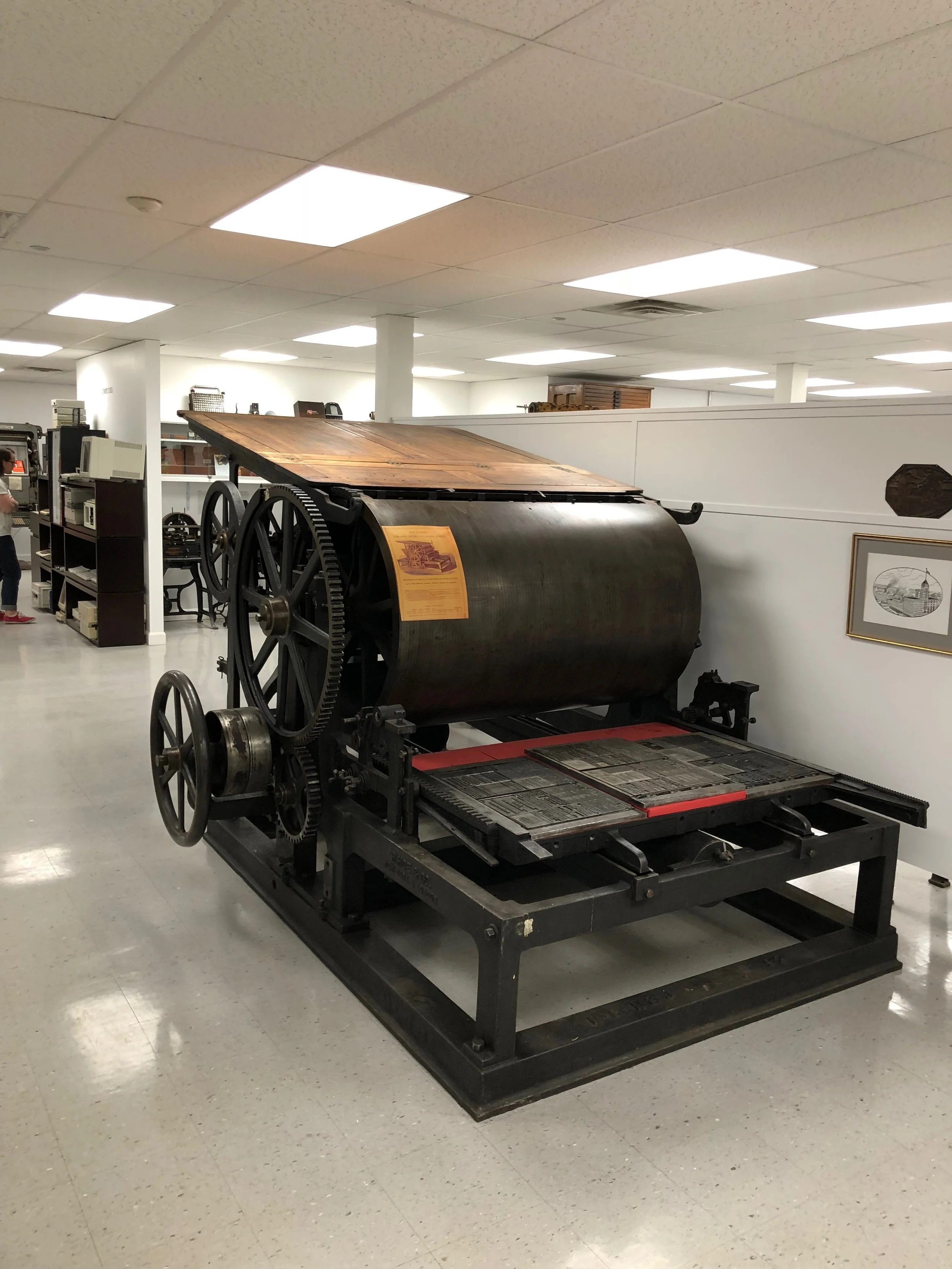 Old large printing press with large gears and a wooden top in a museum setting.
