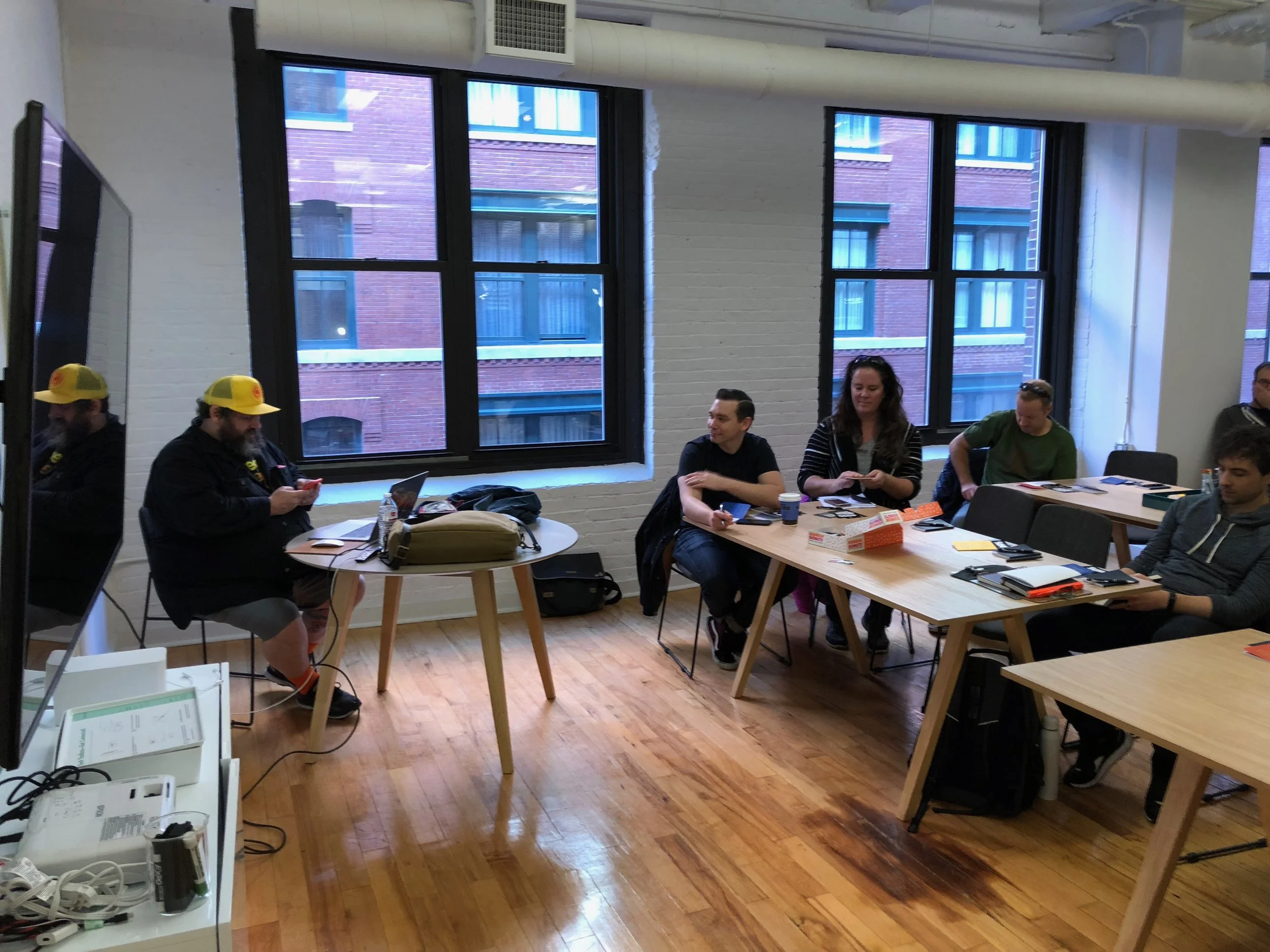 People in a meeting room sitting at tables, some with laptops and notebooks, with a person in a yellow hat on the left and others engaging in conversation or looking at devices.