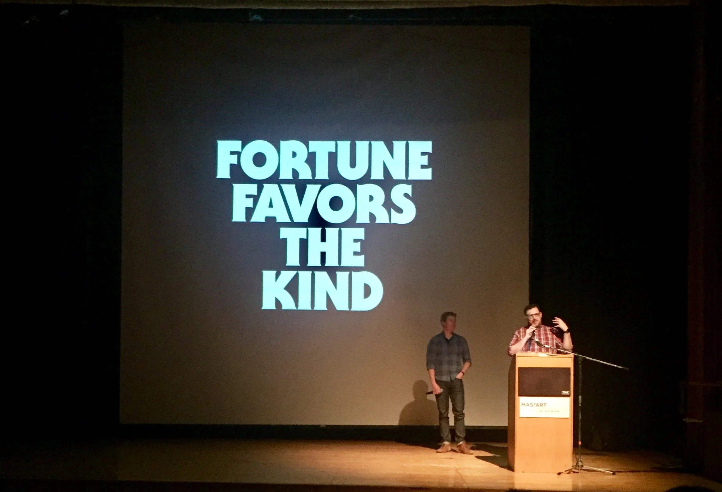 Two men on stage in front of a large screen displaying the words 'Fortune Favors The Kind'. One man is standing behind a podium, speaking into a microphone, while the other stands nearby with hands in pockets.