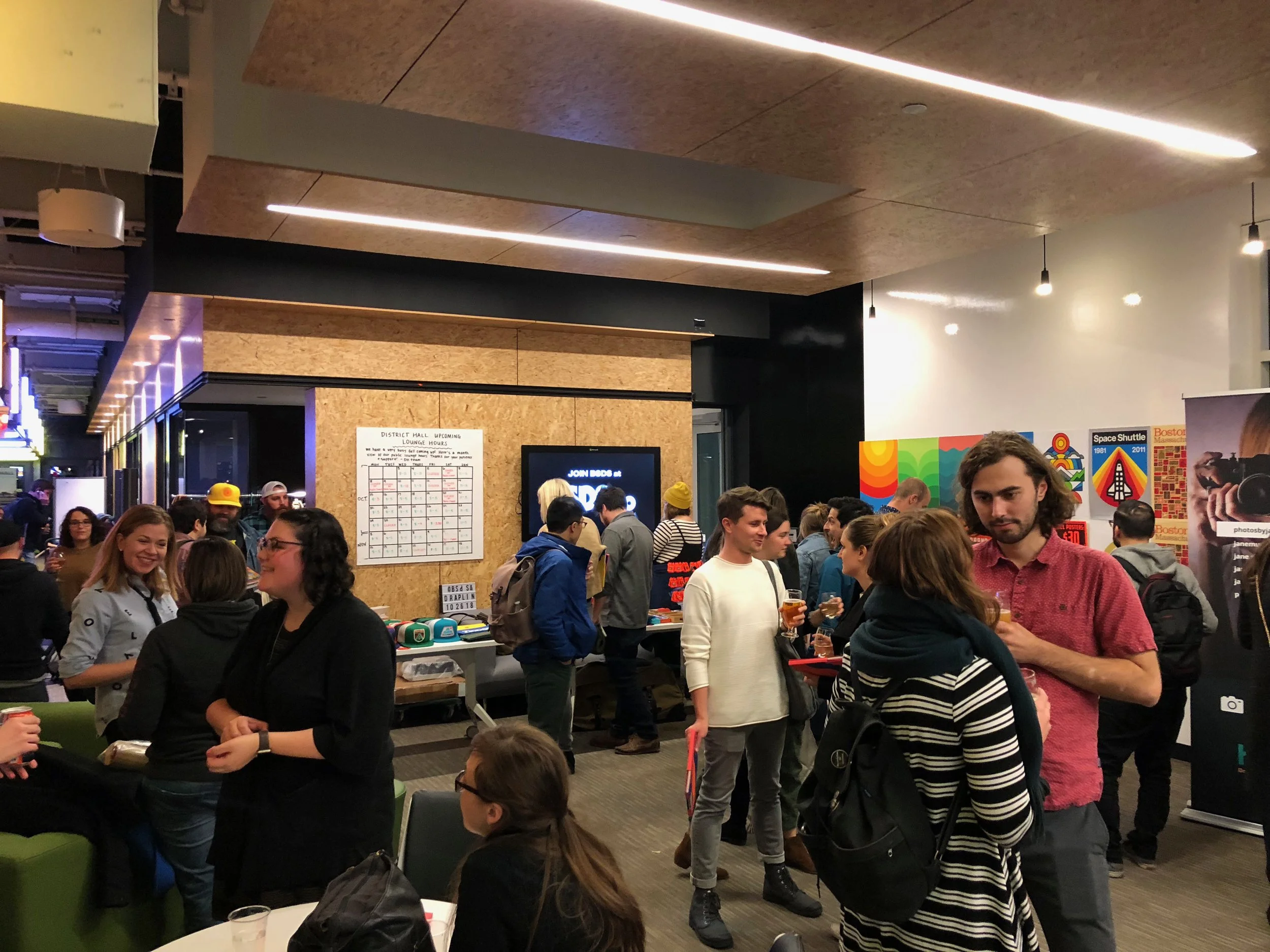 People socializing at an indoor event with colorful posters on the wall, some holding drinks, in a modern space with wood and black decor.