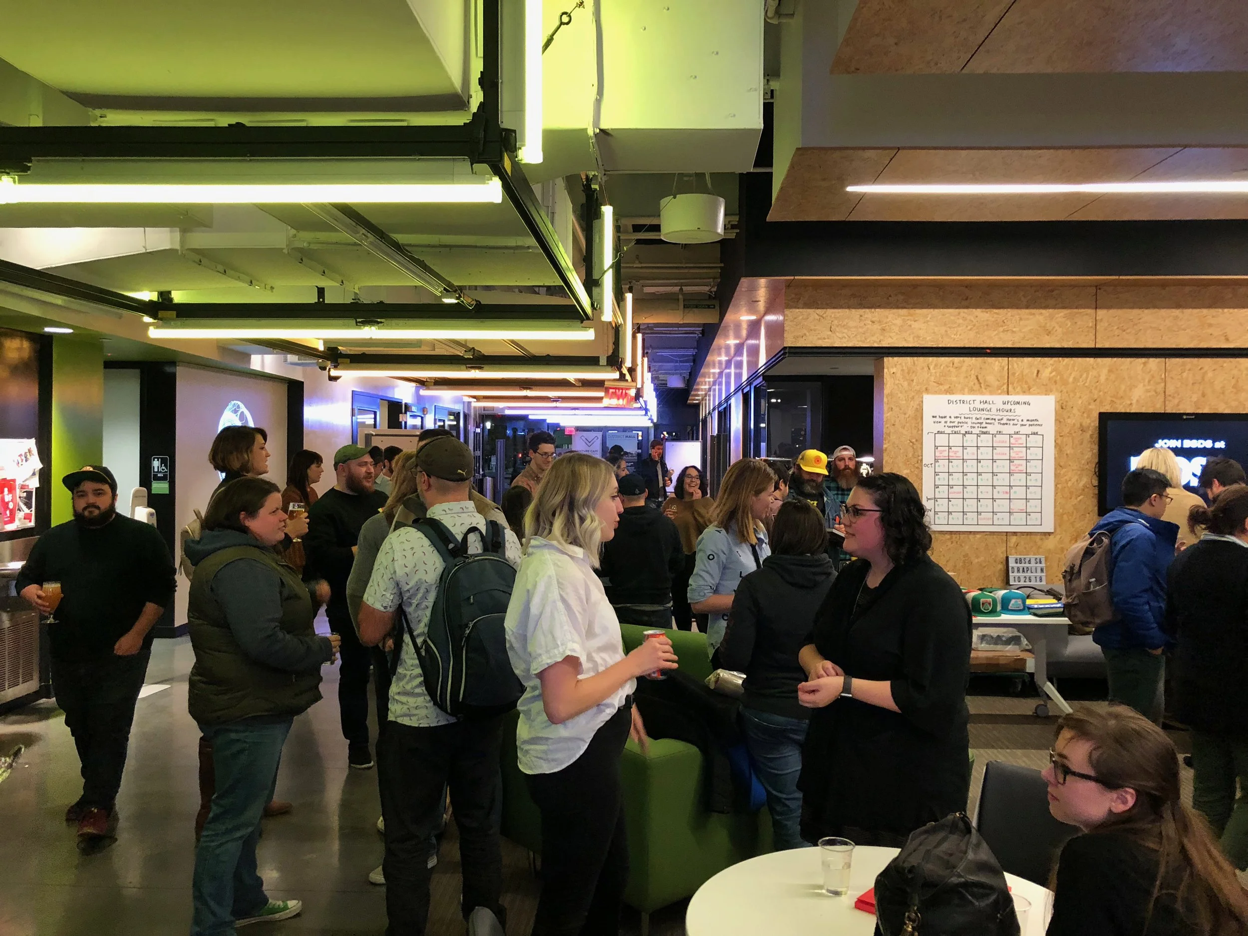 People socializing and standing in line at an indoor event, with some holding drinks, in a modern space with wooden and black accents, bright green lighting, and a large electronic display.