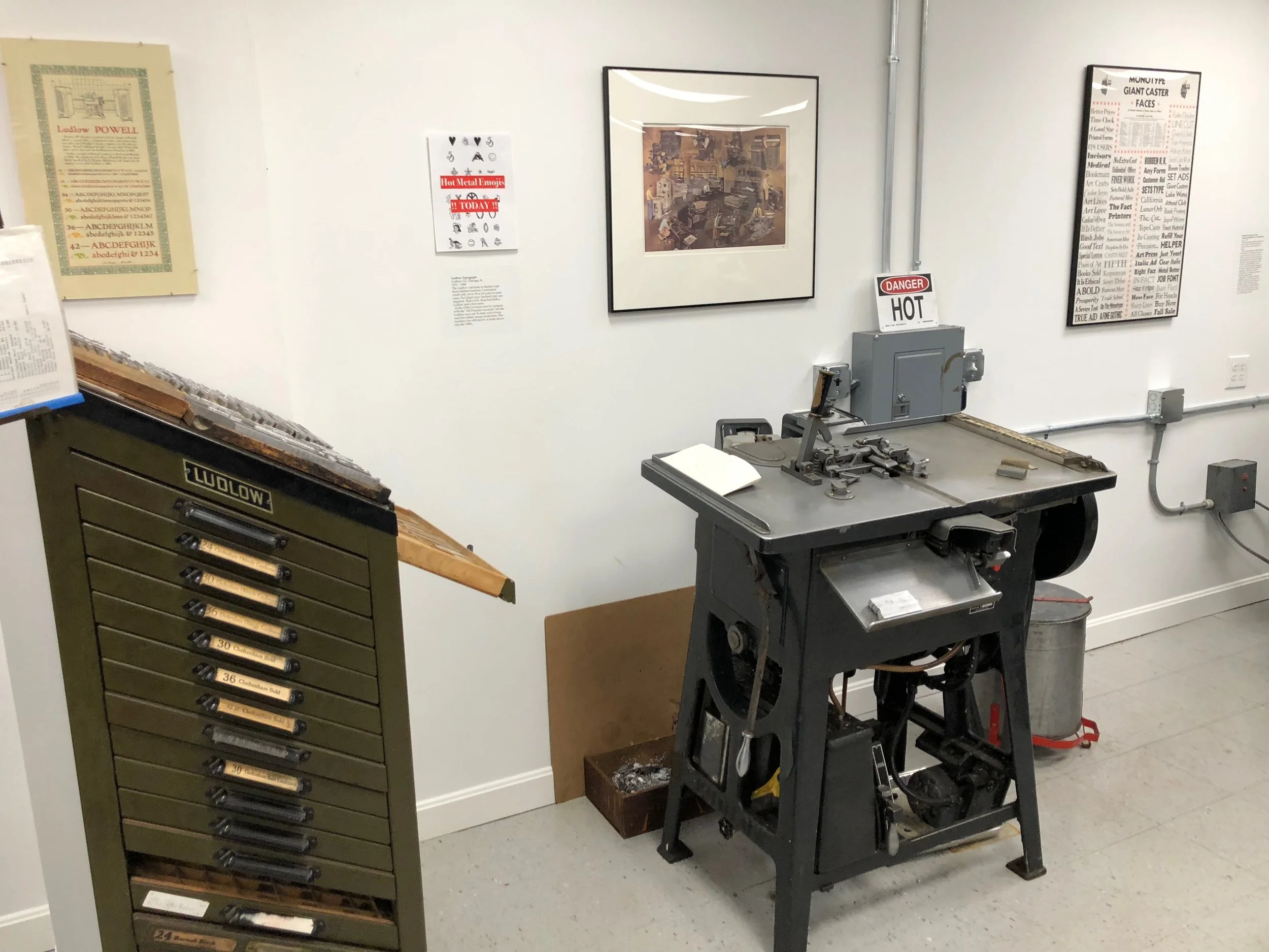 A vintage printing or typesetting workshop with a large metal working machine and a cabinet with labeled drawers for different types or tools. The walls are decorated with informational posters and framed pictures.
