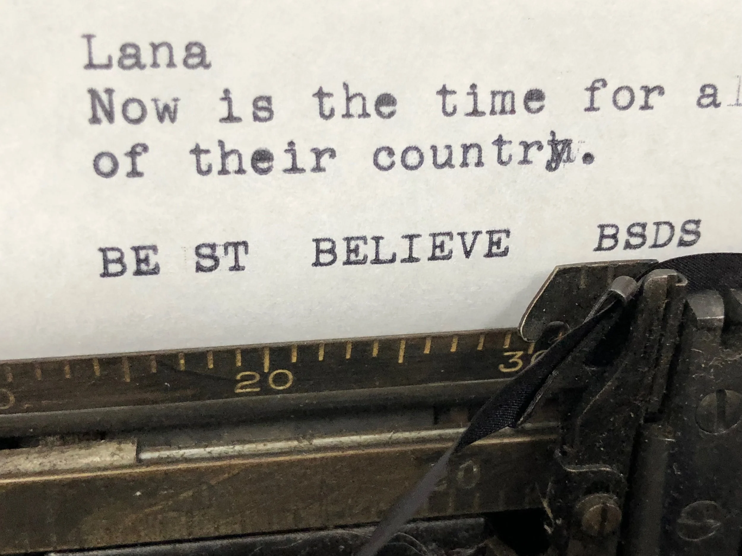 Close-up of a typewriter with a typed note reading 'Lana Now is the time for a of their country. BE ST BELIEVE BSDS'.