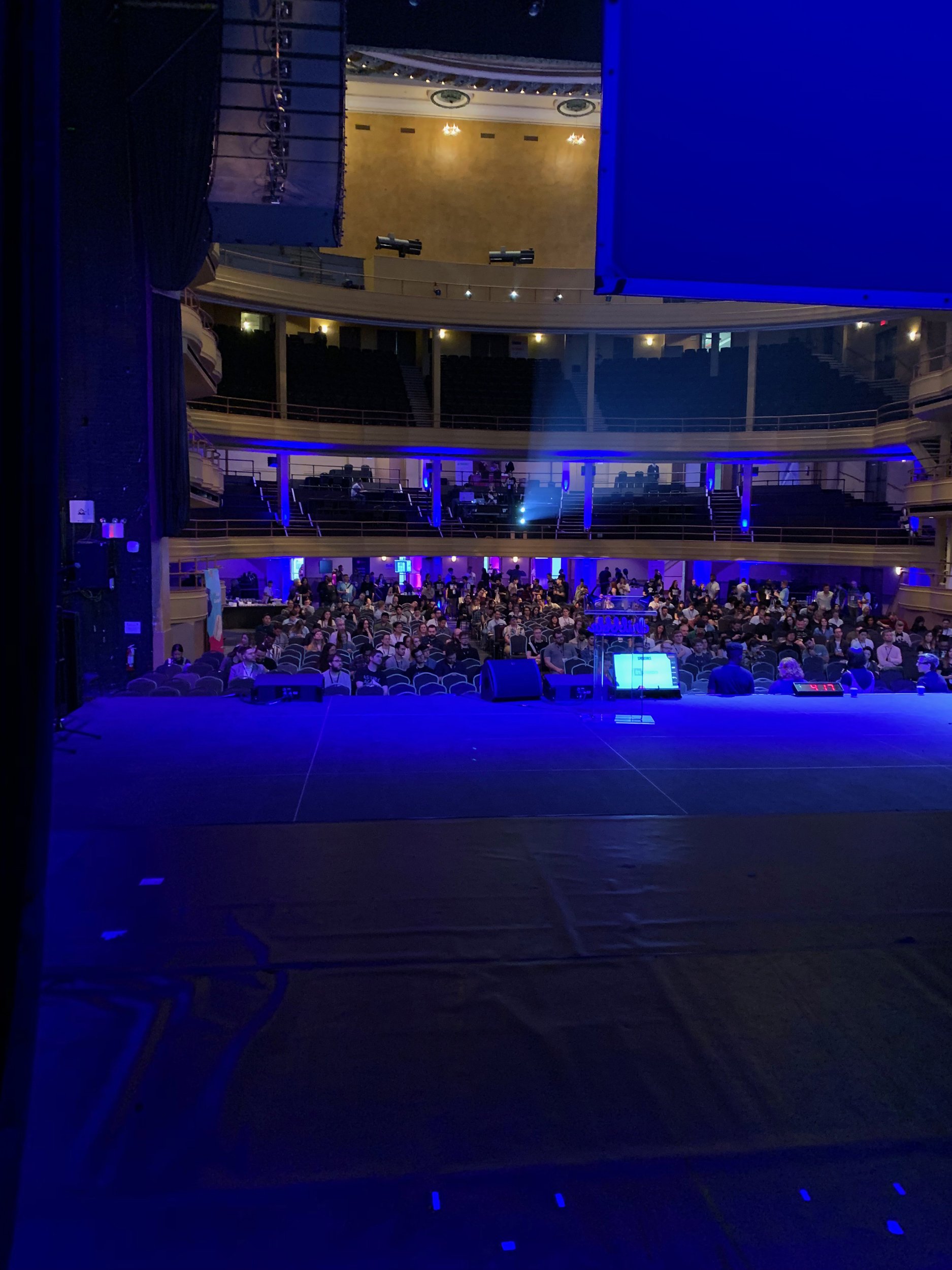 View of a theater or concert hall from the stage, showing rows of audience seating filled with people, with a balcony level above. The room is dimly lit with blue and purple lighting.