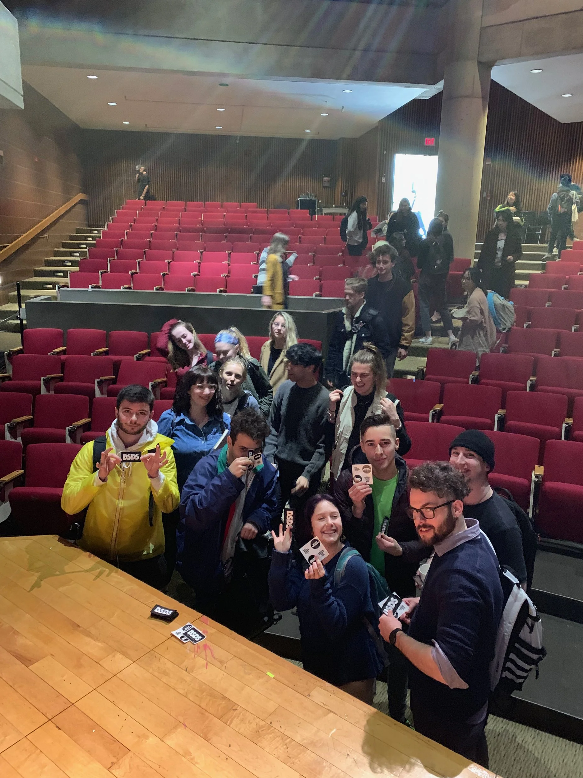 Group of young people smiling and holding tickets in an auditorium with red seats and wooden walls.
