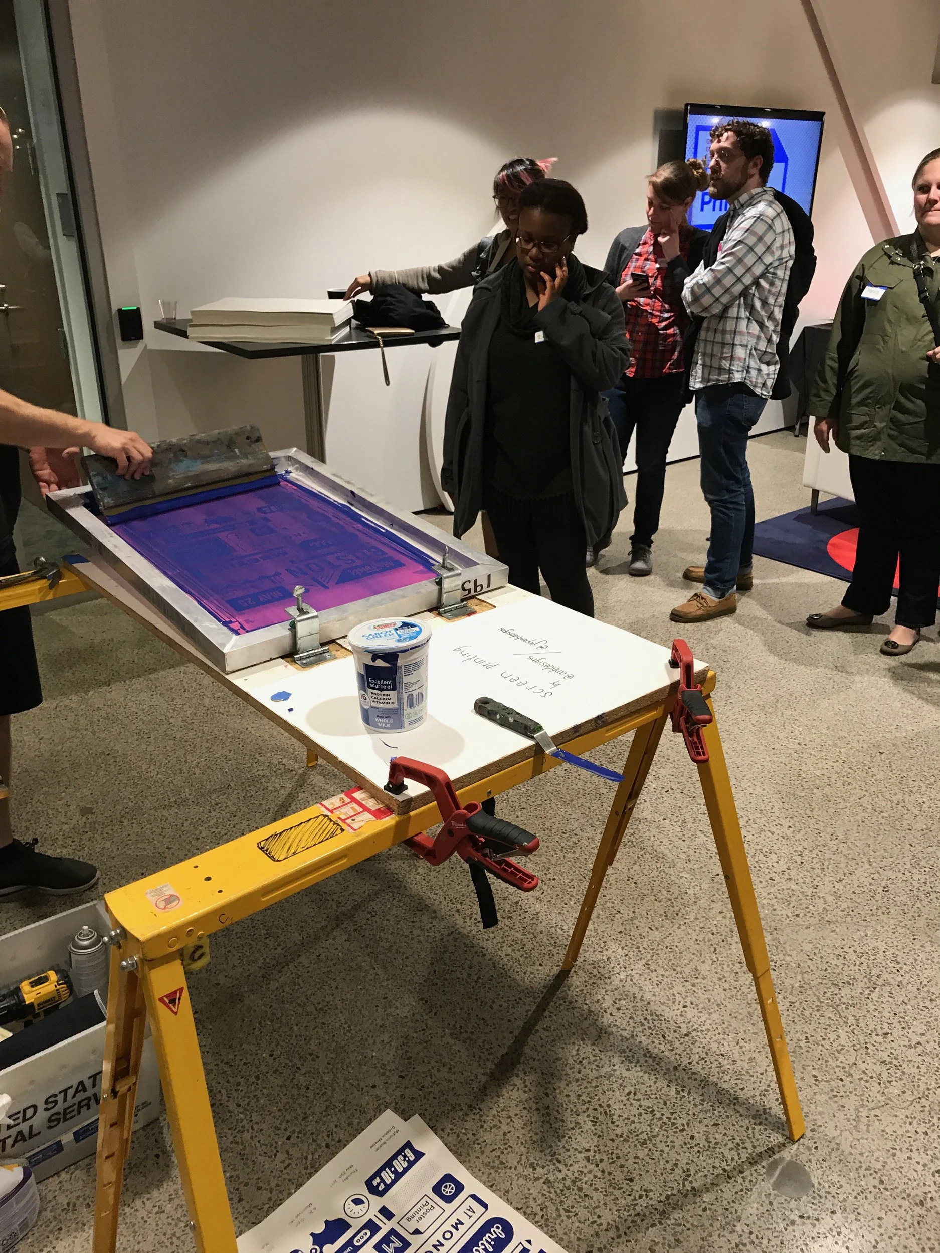 People viewing a printing or printing process in a room with a table and a press.