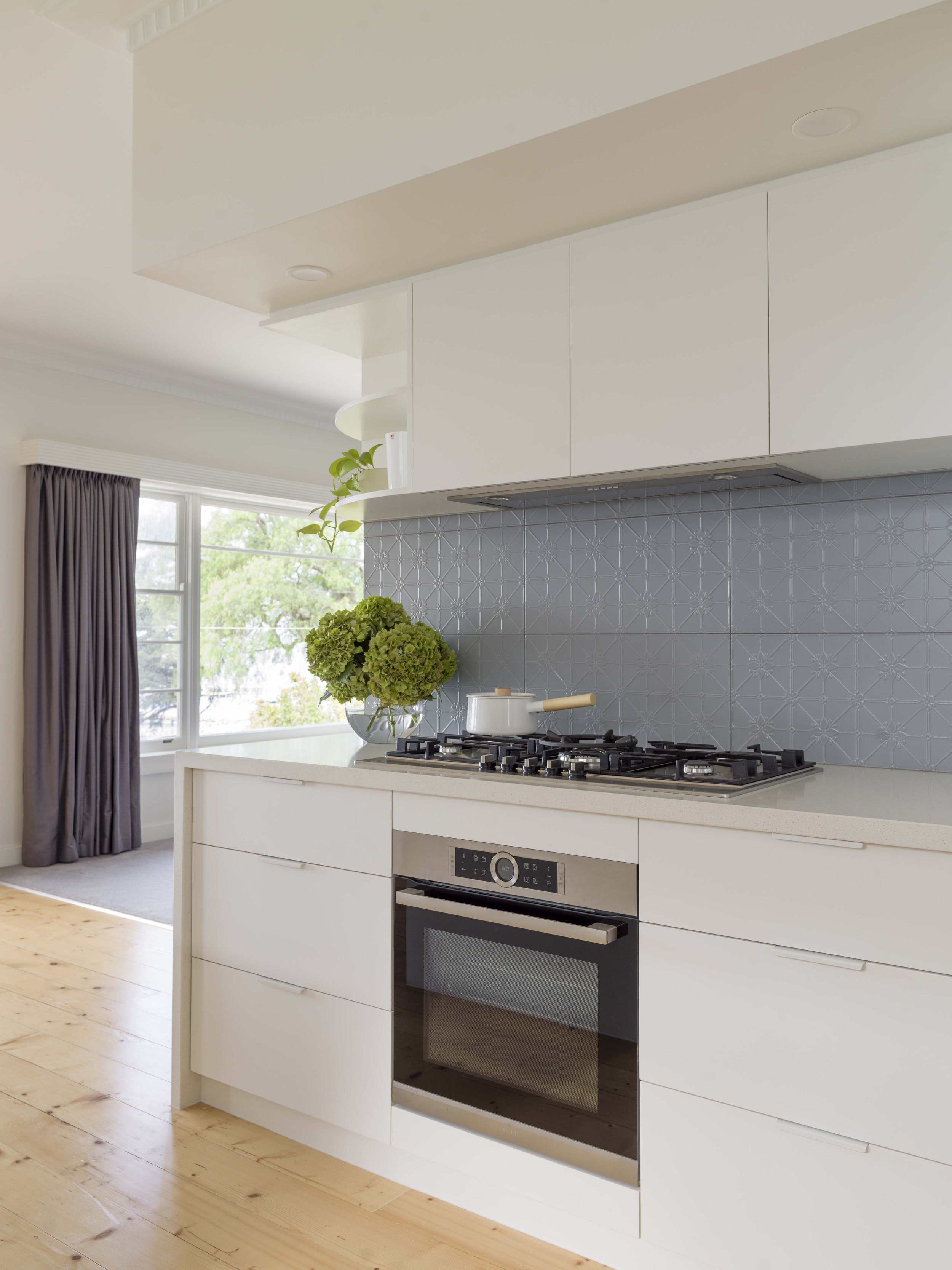 Modern kitchen with white cabinetry, blue patterned tile backsplash, stainless steel oven, gas stove, white countertop with a white pot, green hydrangea flowers in a glass vase, window with dark curtains, and hanging plants.