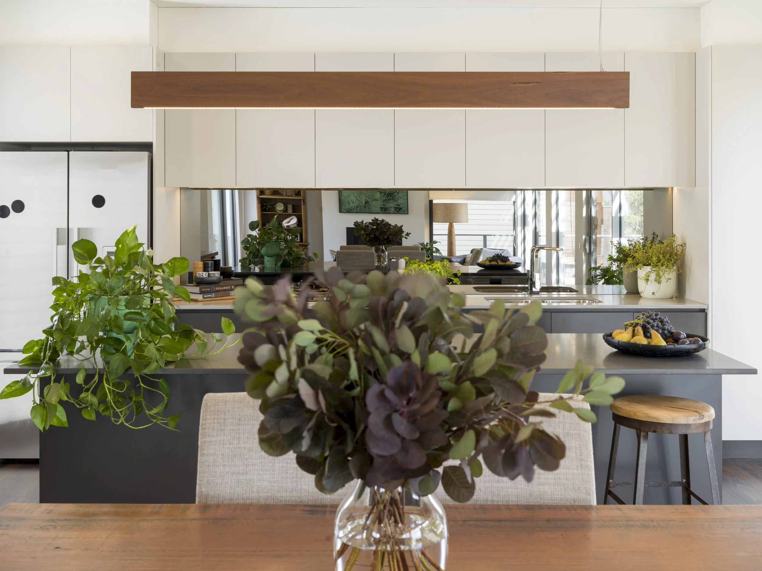 A view of a modern, minimalist kitchen and living room, seen through a glass vase with green and purple houseplants in the foreground on a wooden table.