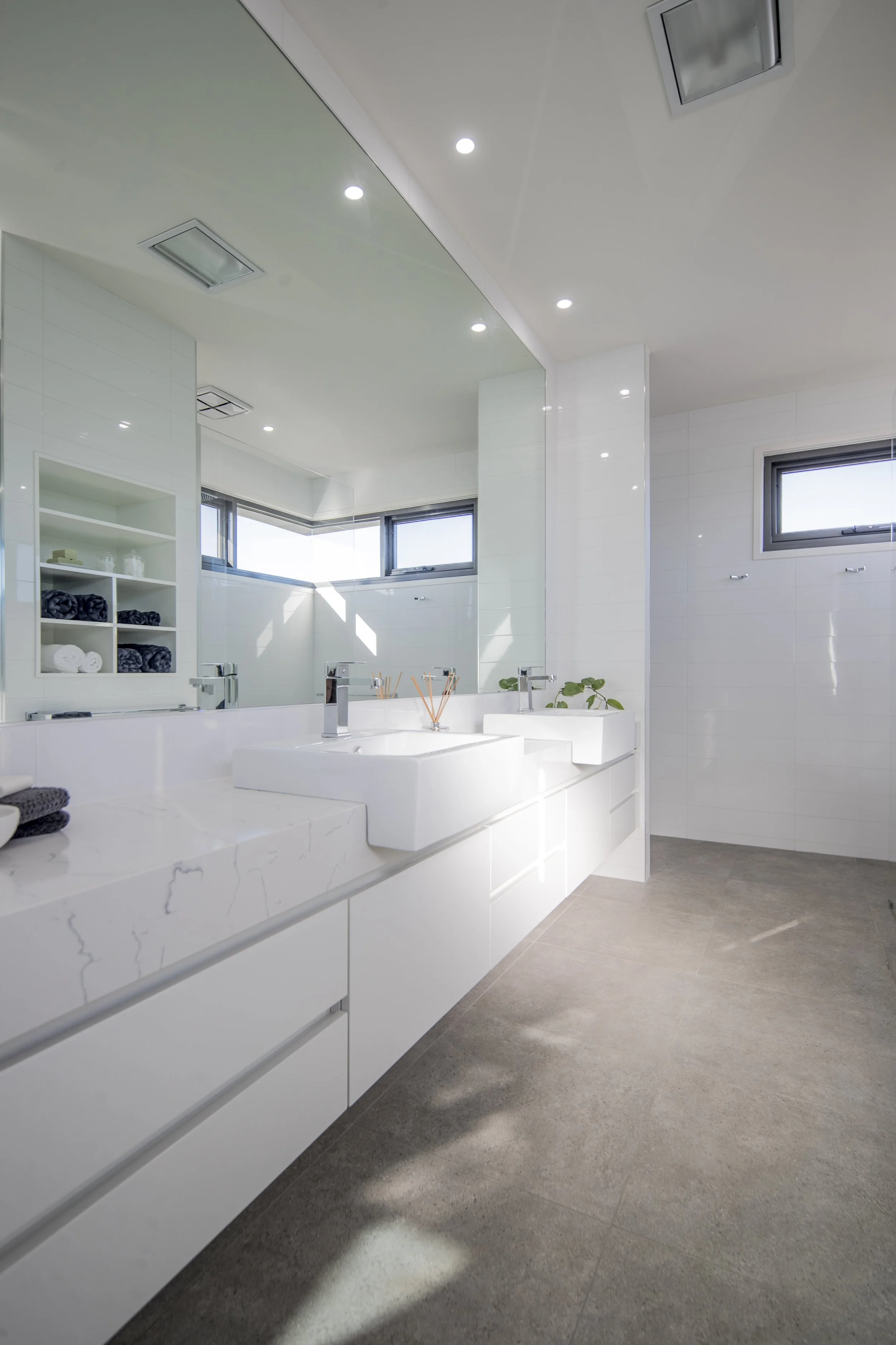 Modern bathroom with white vanity, double sinks, large mirror, and grey tiled floor. There are high horizontal windows, ceiling recessed lights, and a small plant on the counter.