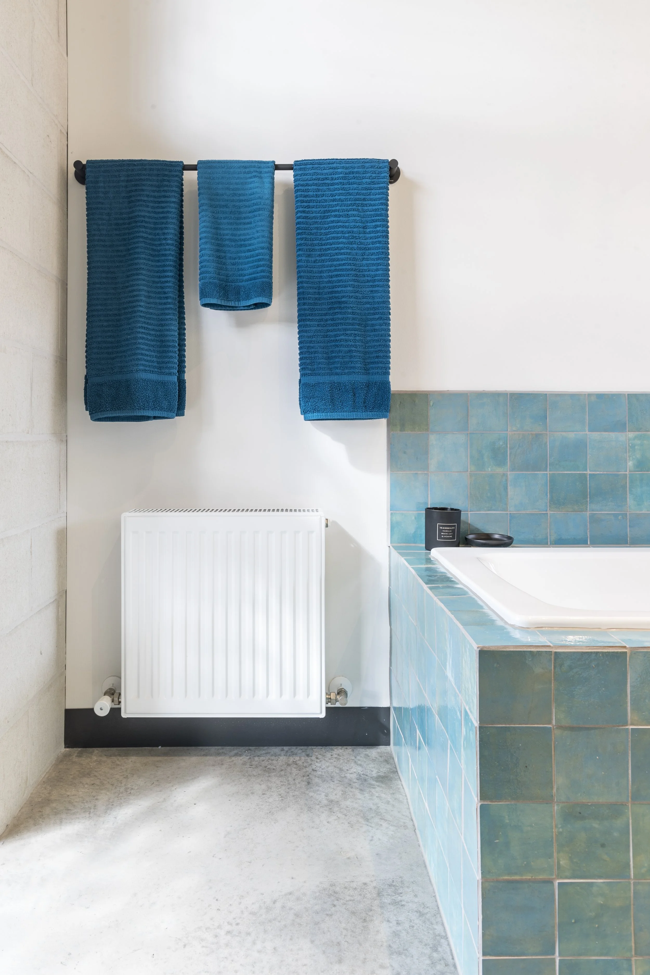 Bathroom with blue towels hanging on a black towel rack, white wall, radiators below, part of a tub with teal tile surround, and a small black container on the tub ledge.