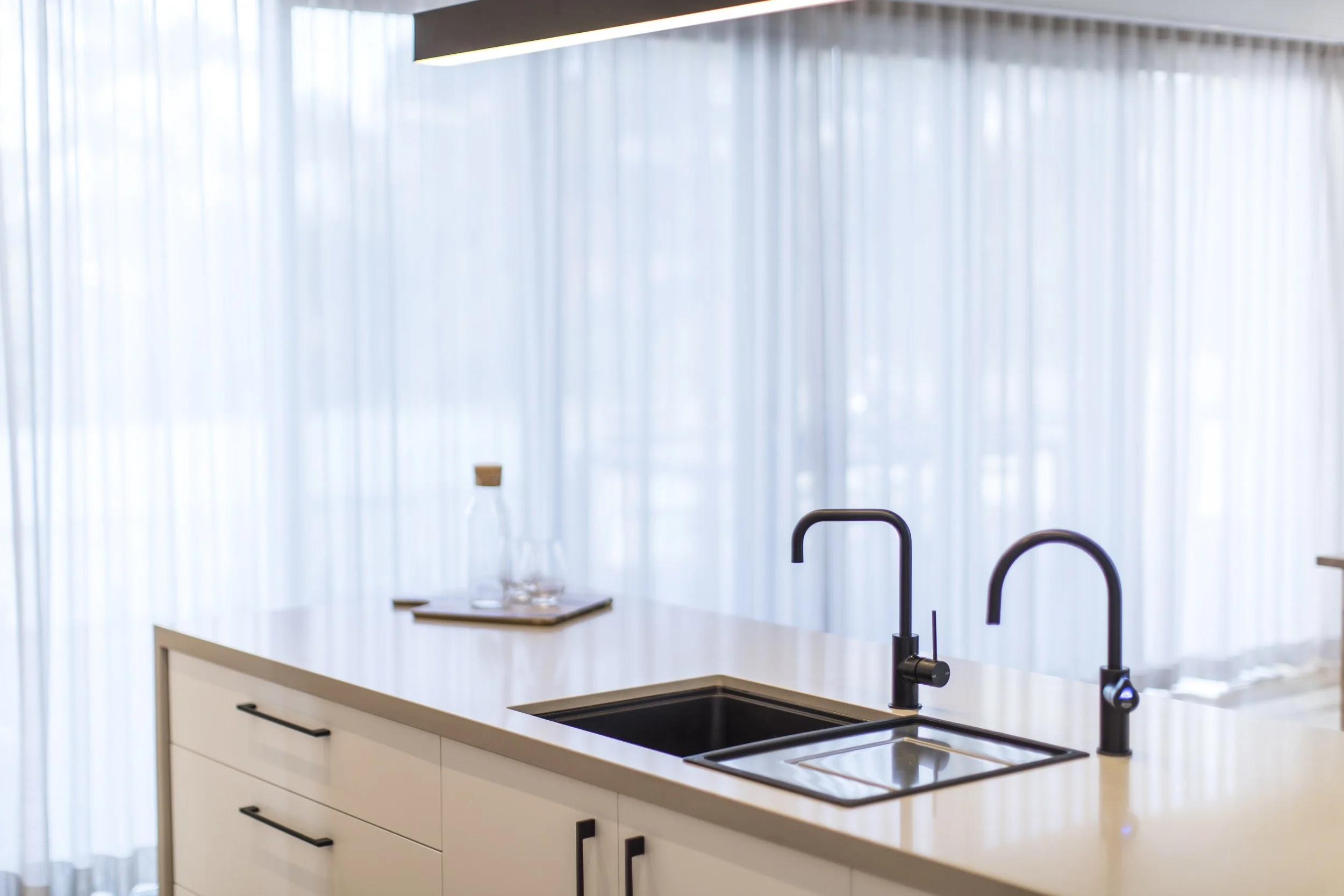 Modern kitchen with a white island, black sink, and black faucet, with a sheer curtain and a ledge holding glassware in the background.
