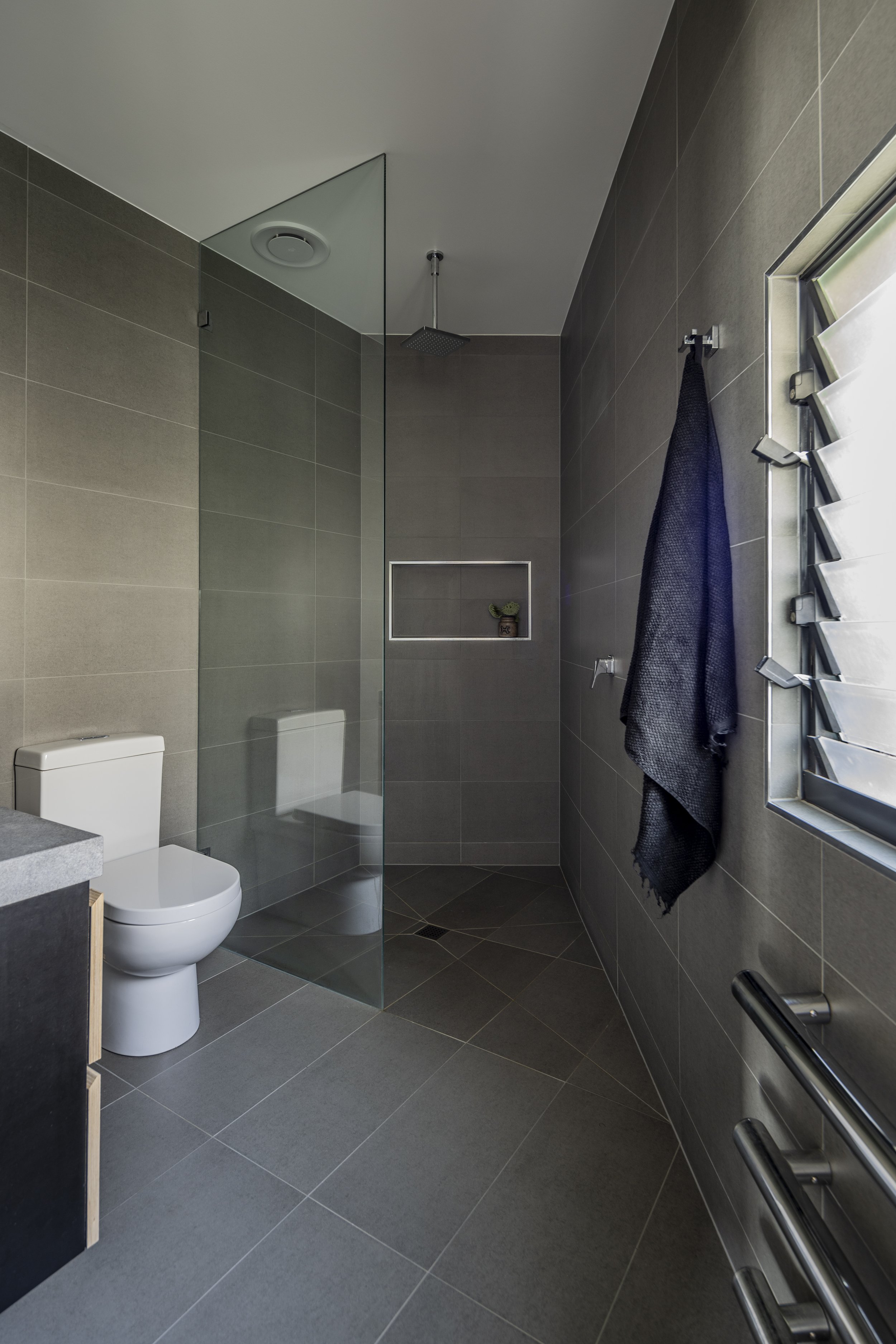 Modern bathroom with gray floor and wall tiles, a toilet, a glass shower wall with a rain showerhead, a black towel hanging on a rack, and a window with adjustable blinds.
