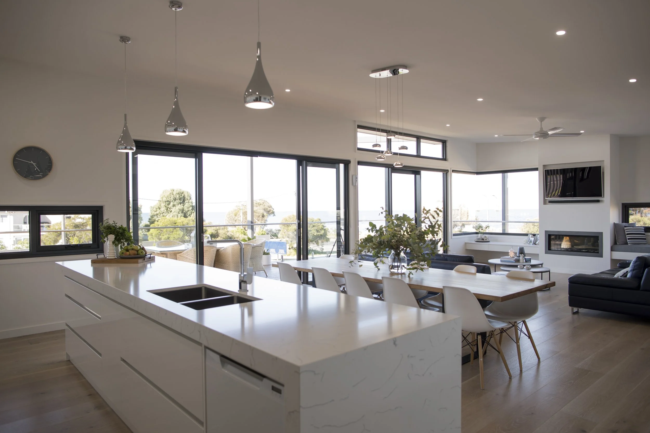 Modern open-concept living room and kitchen with large windows, a white kitchen island, a dining table with white chairs, a black sofa, and a wall-mounted TV. Several pendant lights hang from the ceiling, and a ceiling fan is installed. There is a wa